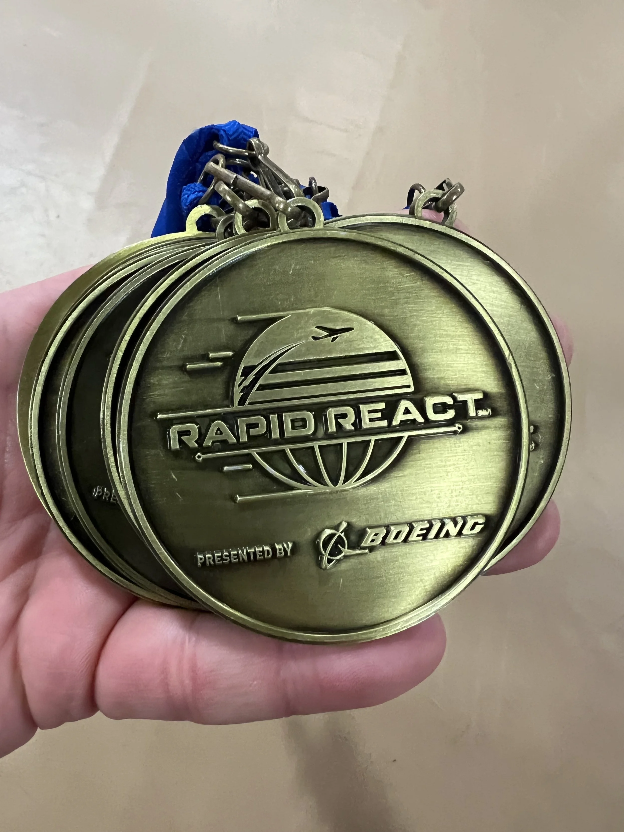 A person is holding a large metallic medal with the logo and text for 'Rapid React,' presented by Boeing, featuring a stylized globe and swooping lines.