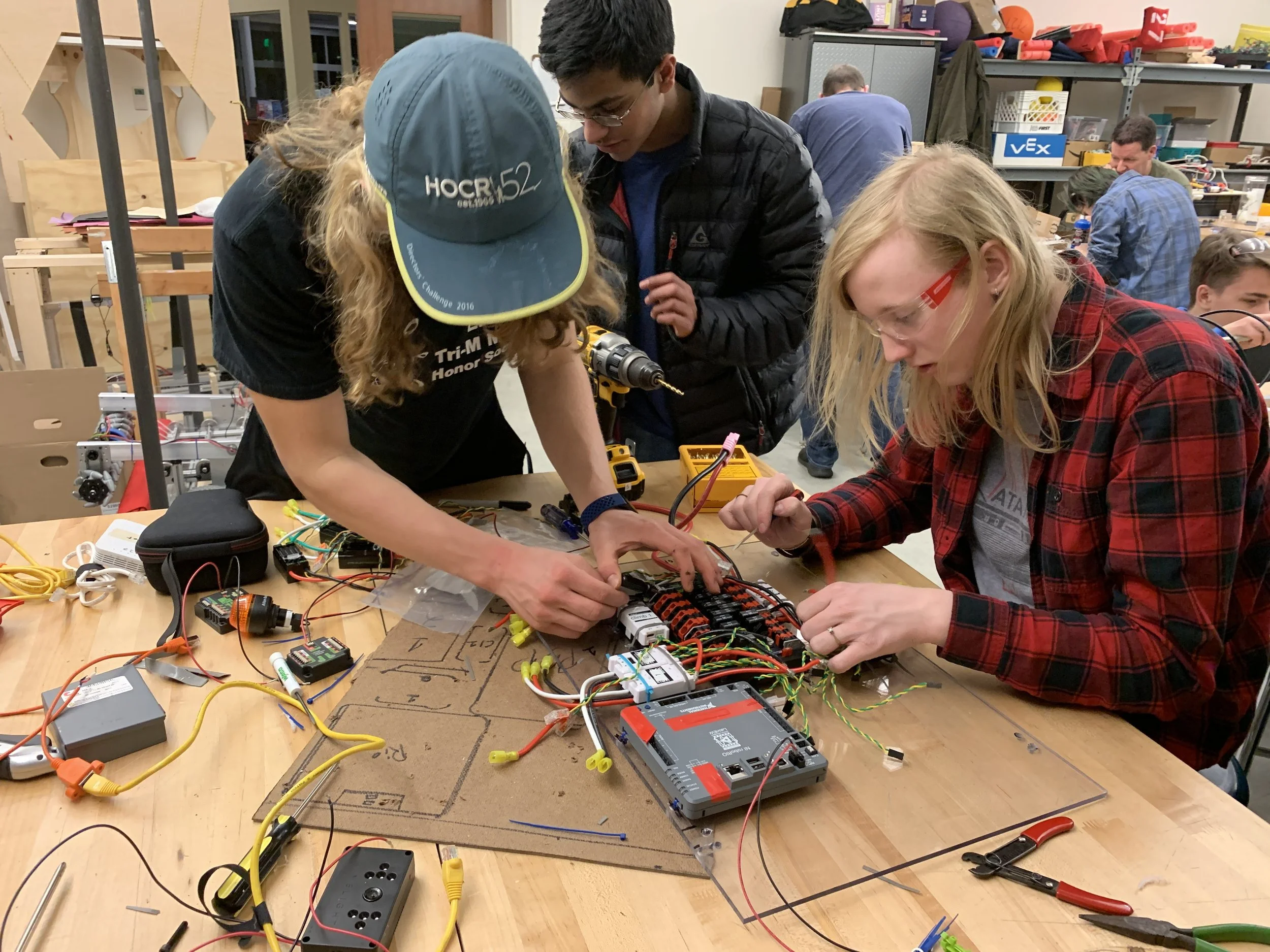 Several young people working together on an electronics project at a workshop table, with tools, wires, and electronic components.