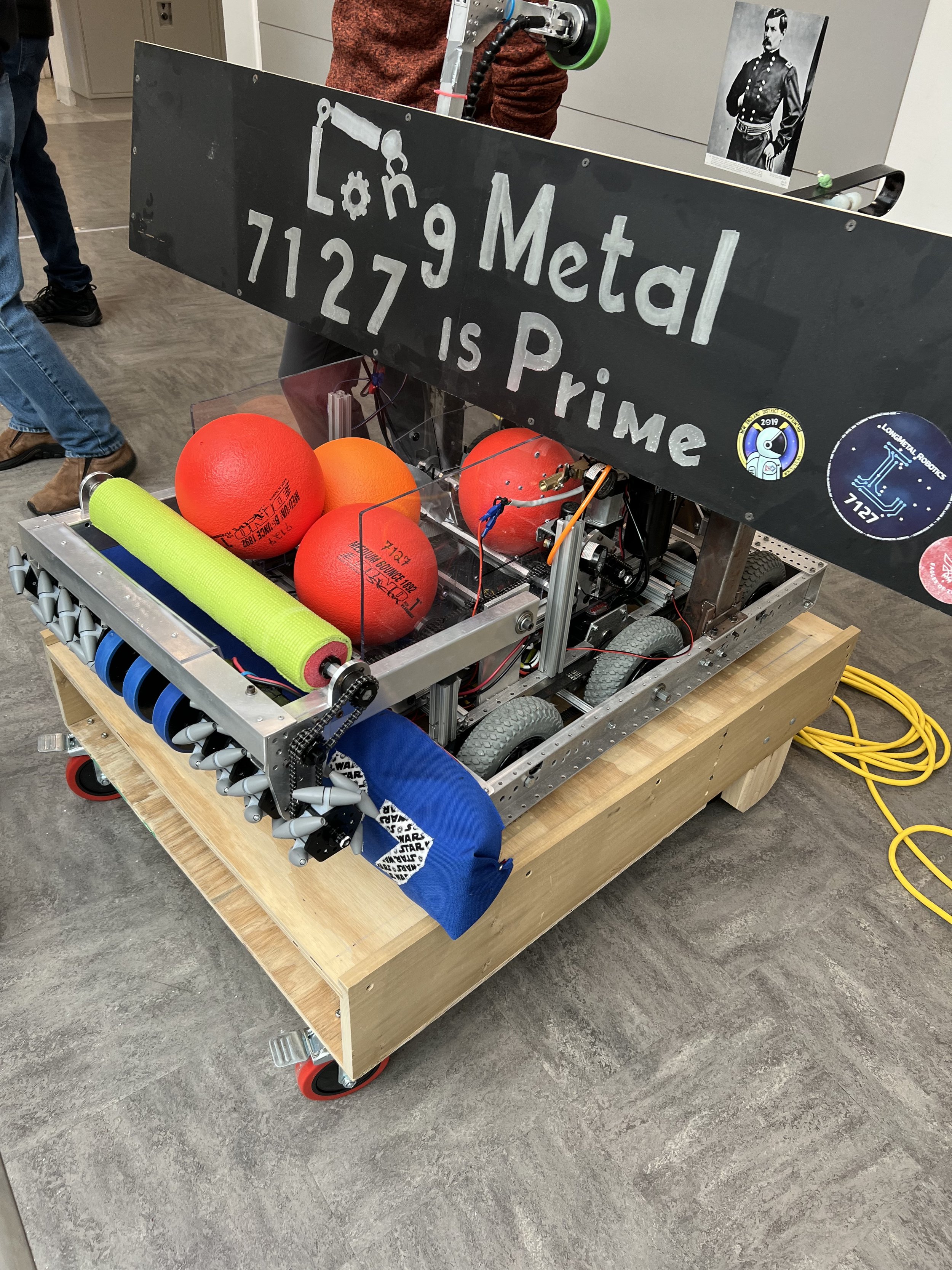 A robot built for a science competition, with a sign that reads 'Lone Metal 7127 is Prime', equipped with a green foam roller, red balls, and various mechanical parts, set on a wooden platform and surrounded by wires and people in the background.