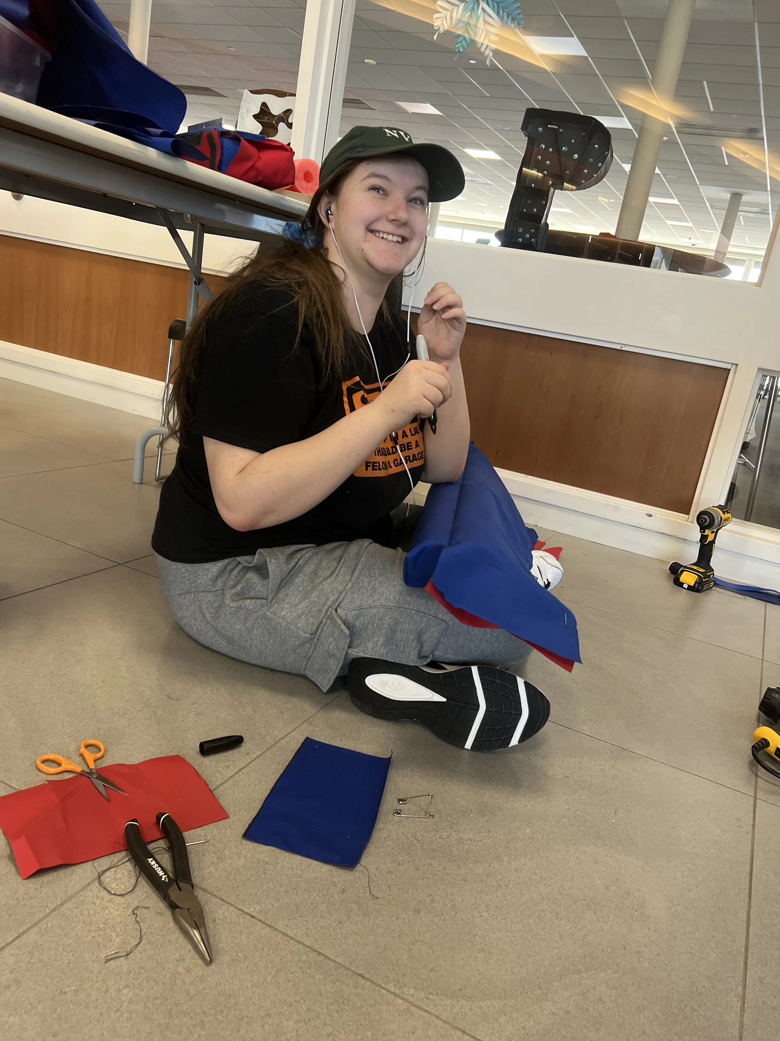 A young woman sitting on the floor at a craft or activity station, smiling, wearing a cap and headphones, surrounded by scissors, fabric, and tools, with a table behind her and a window reflecting the ceiling lights.