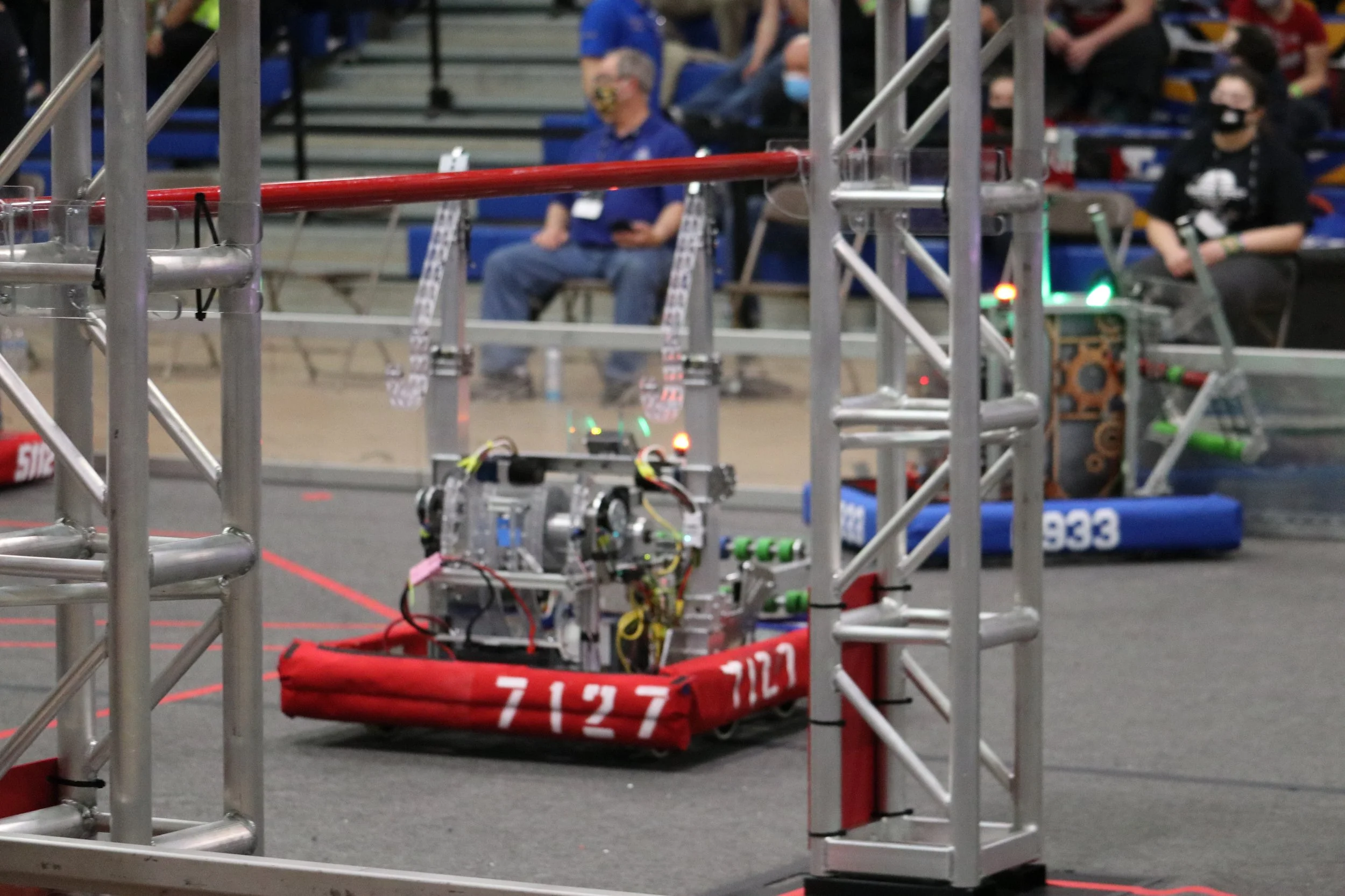 A robotics competition with two robots on the field, surrounded by metal barriers and spectators in the background, some wearing masks.