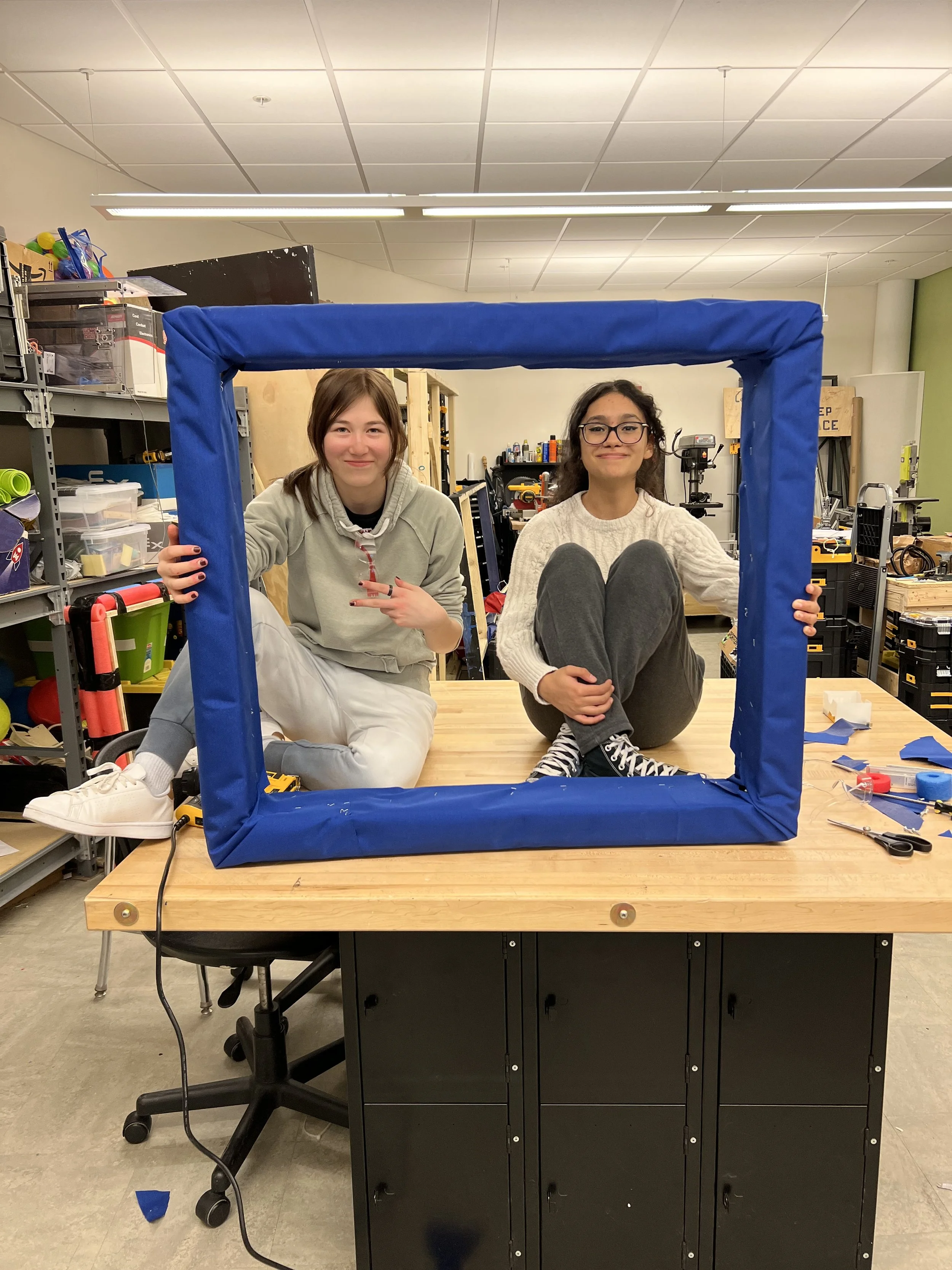 Two women sitting on a workshop table behind a blue fabric-wrapped frame, inside a workshop with shelves of tools and materials.