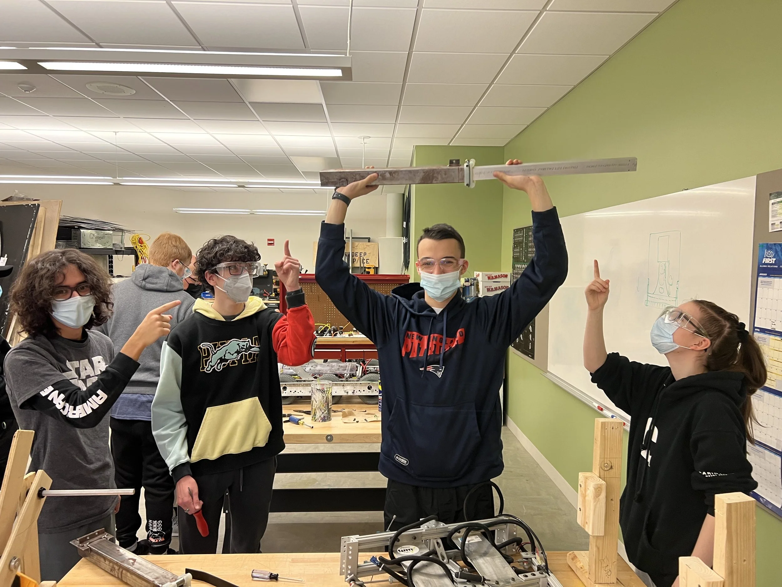 A group of five students wearing safety goggles and face masks in a classroom workshop, with three of them pointing at a large ruler held by a student in the center, who is lifting it overhead. The classroom has green and white walls, workbenches, an
