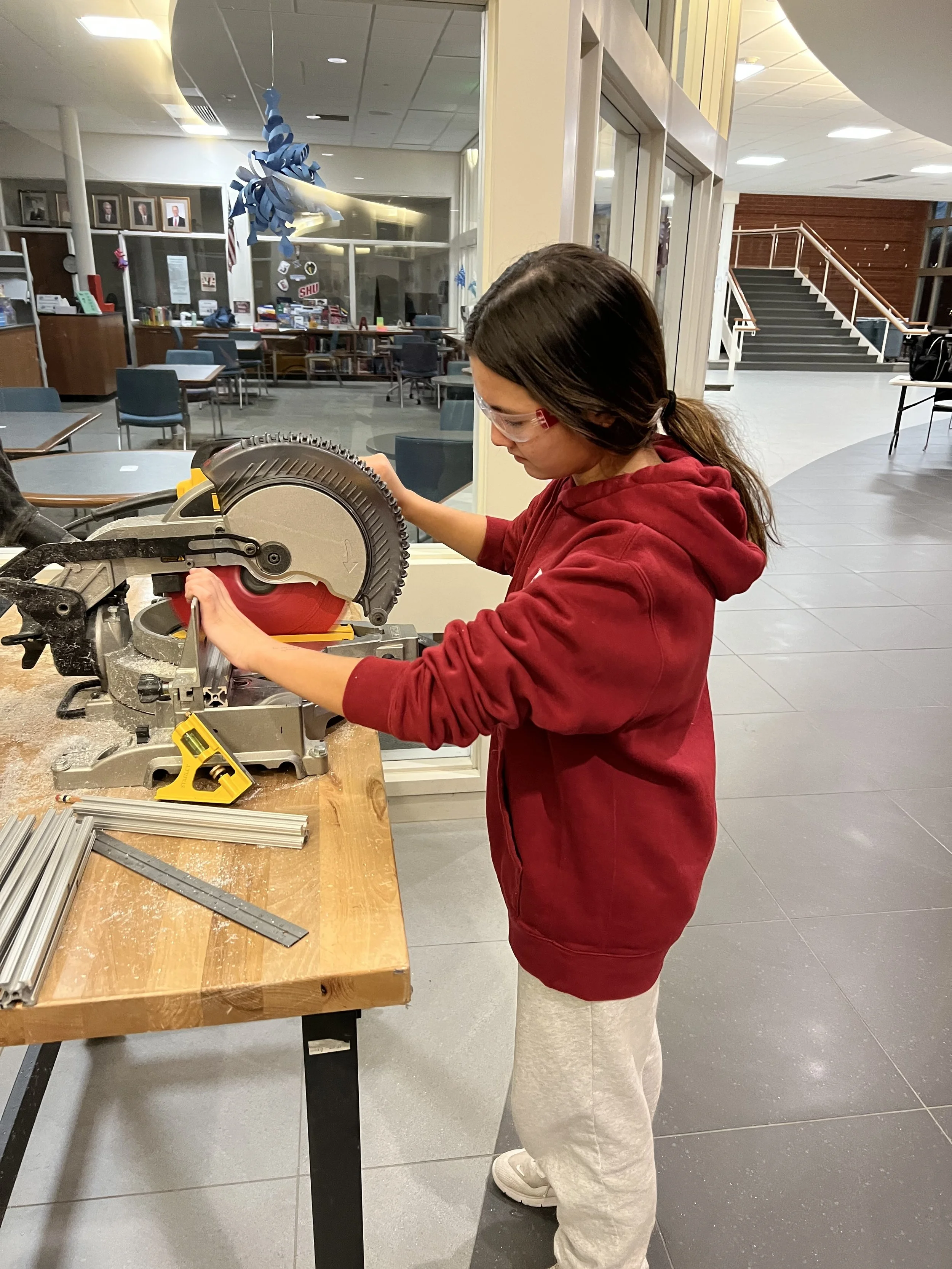 A girl wearing safety glasses and a red hoodie using a miter saw on a workbench with metal pieces in a workshop or classroom setting.