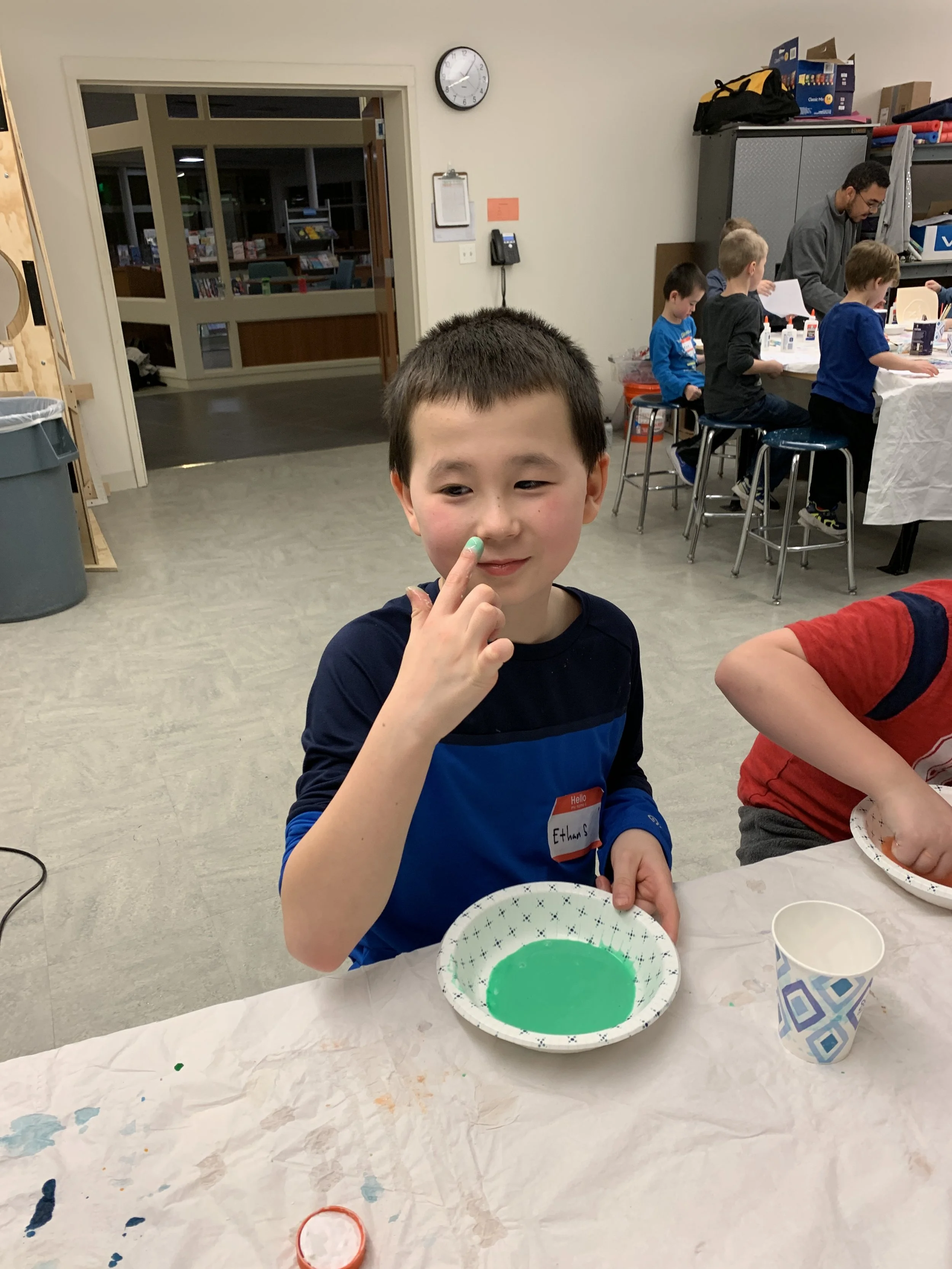 A young boy at a table with green paint on his finger, holding a paper plate with green paint, overlapping with other children and adults in the background in an indoor classroom or activity space.