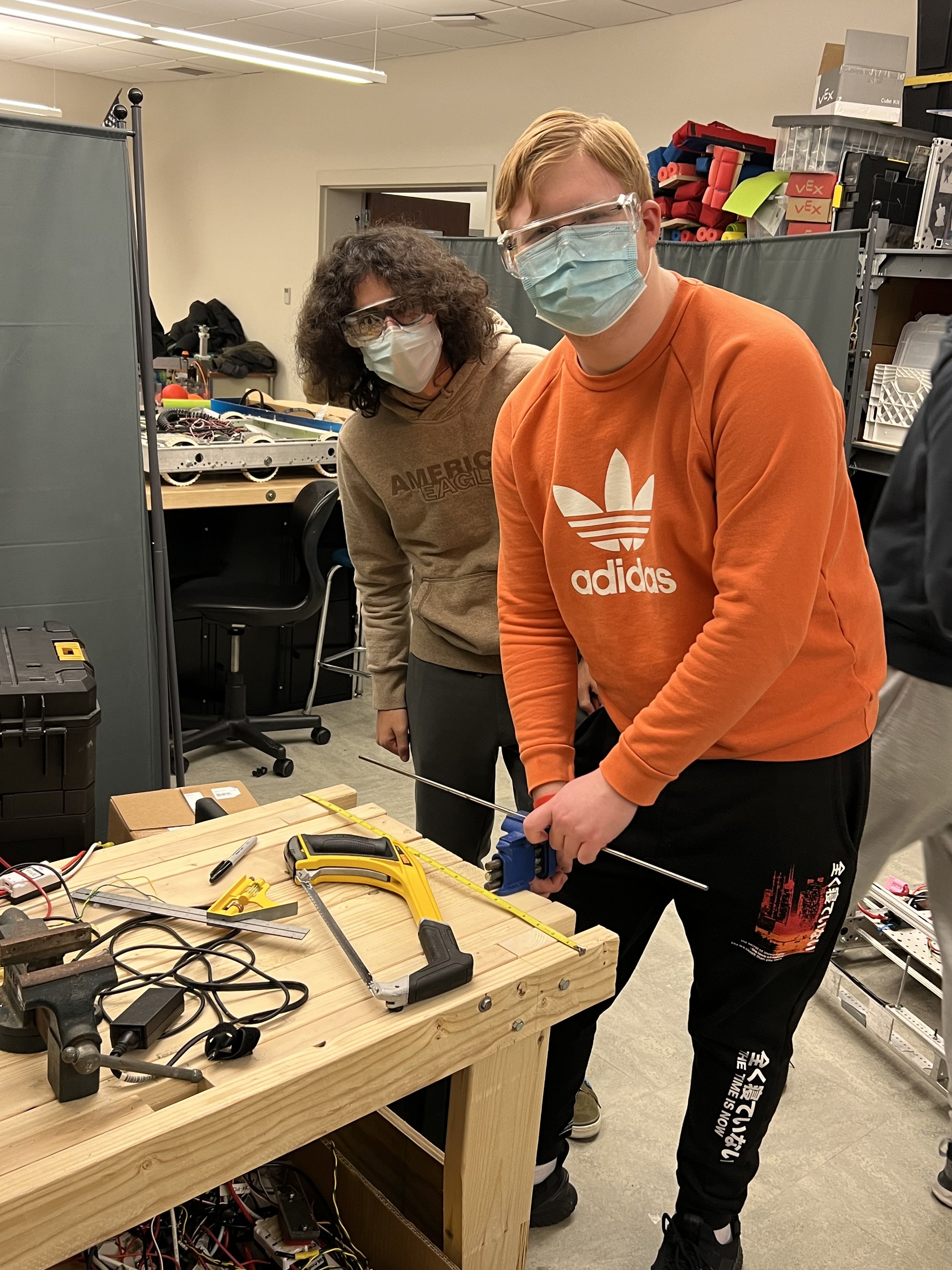 Two individuals wearing masks and safety goggles working on a project involving a long metal rod in a workshop setting with tools and electronic equipment on a wooden workbench.