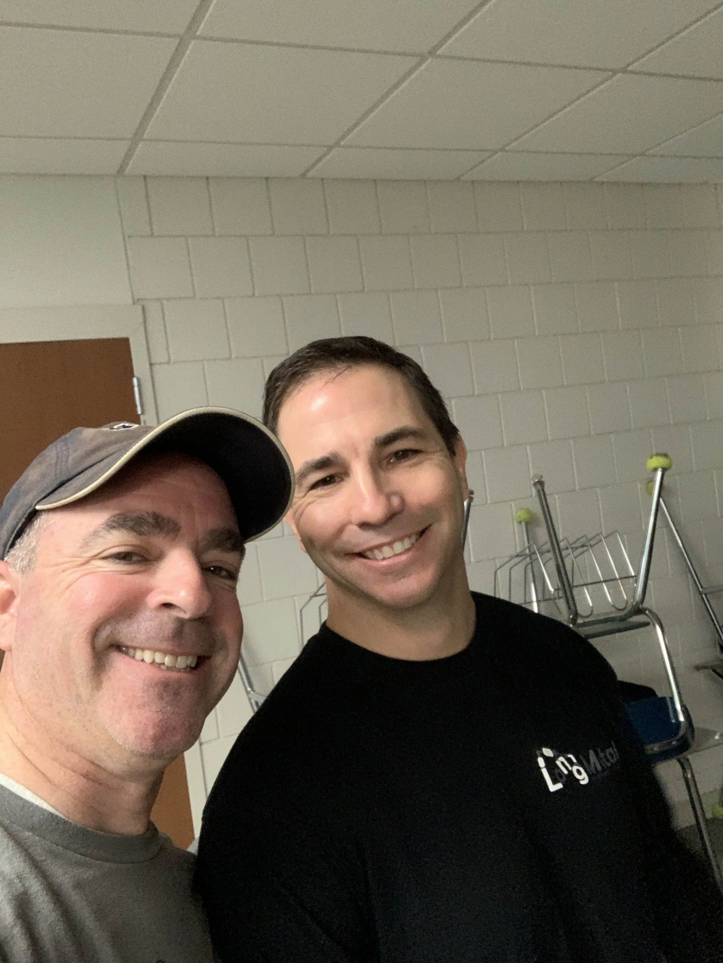 Two men smiling for a selfie indoors, with a background of a white brick wall and stacked chairs.