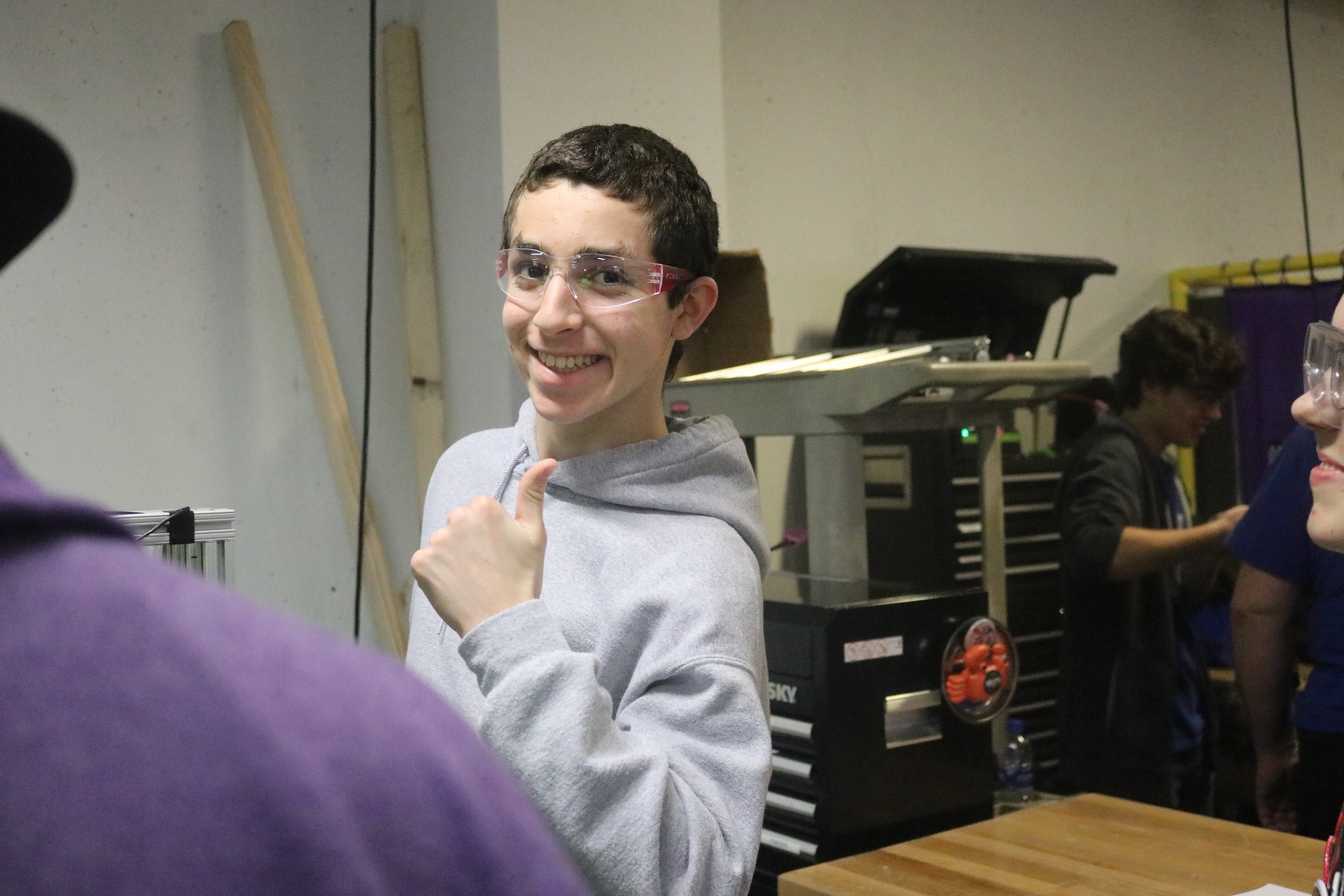 A young man with short dark hair, wearing safety glasses and a gray hoodie, smiling and giving a thumbs-up in a workshop or classroom setting, with a black electronic device and another person in the background.