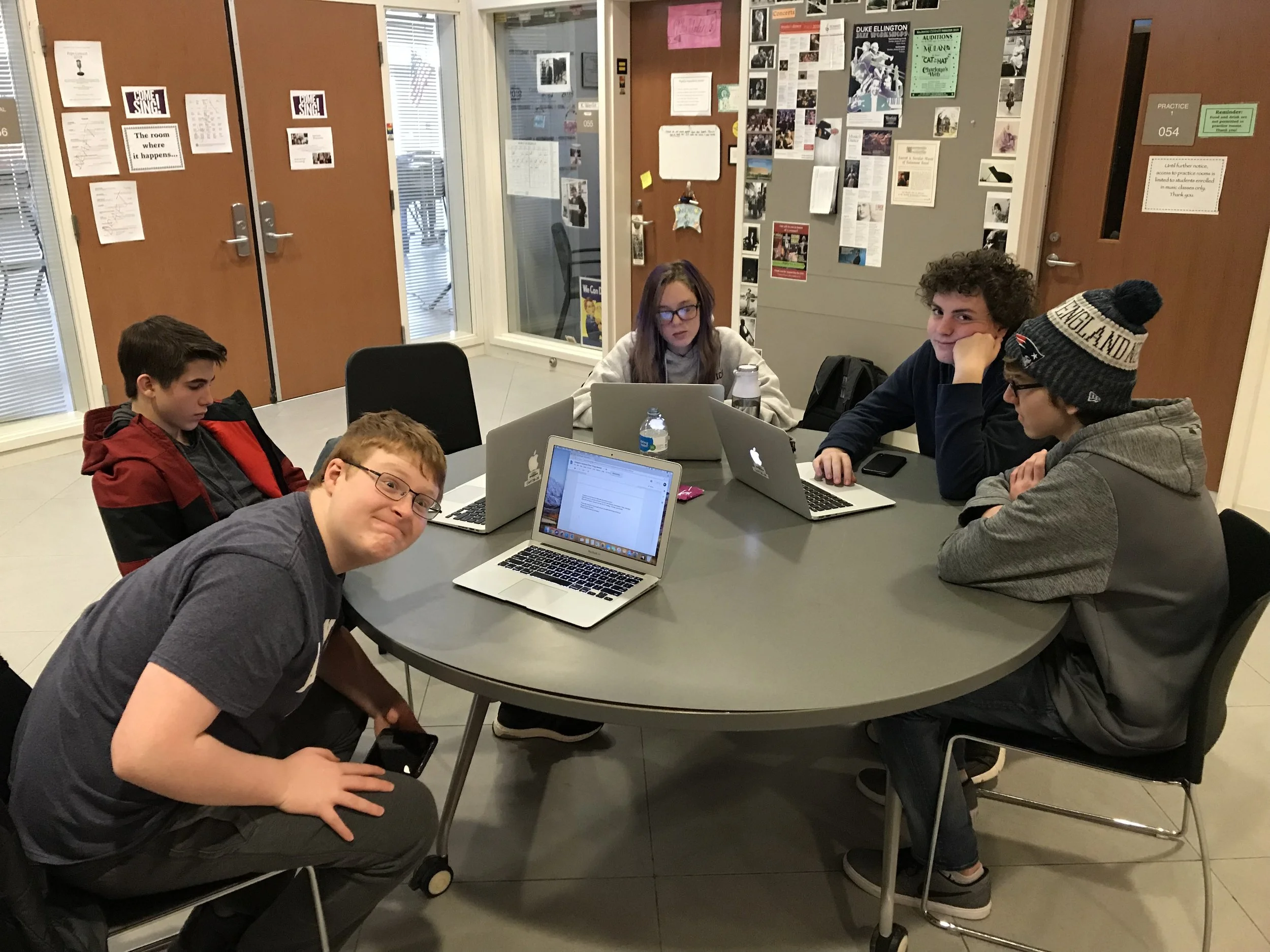 Six teenagers sitting around a gray oval table with laptops, some looking at the camera. The background has wooden doors and bulletin boards with flyers and photos.