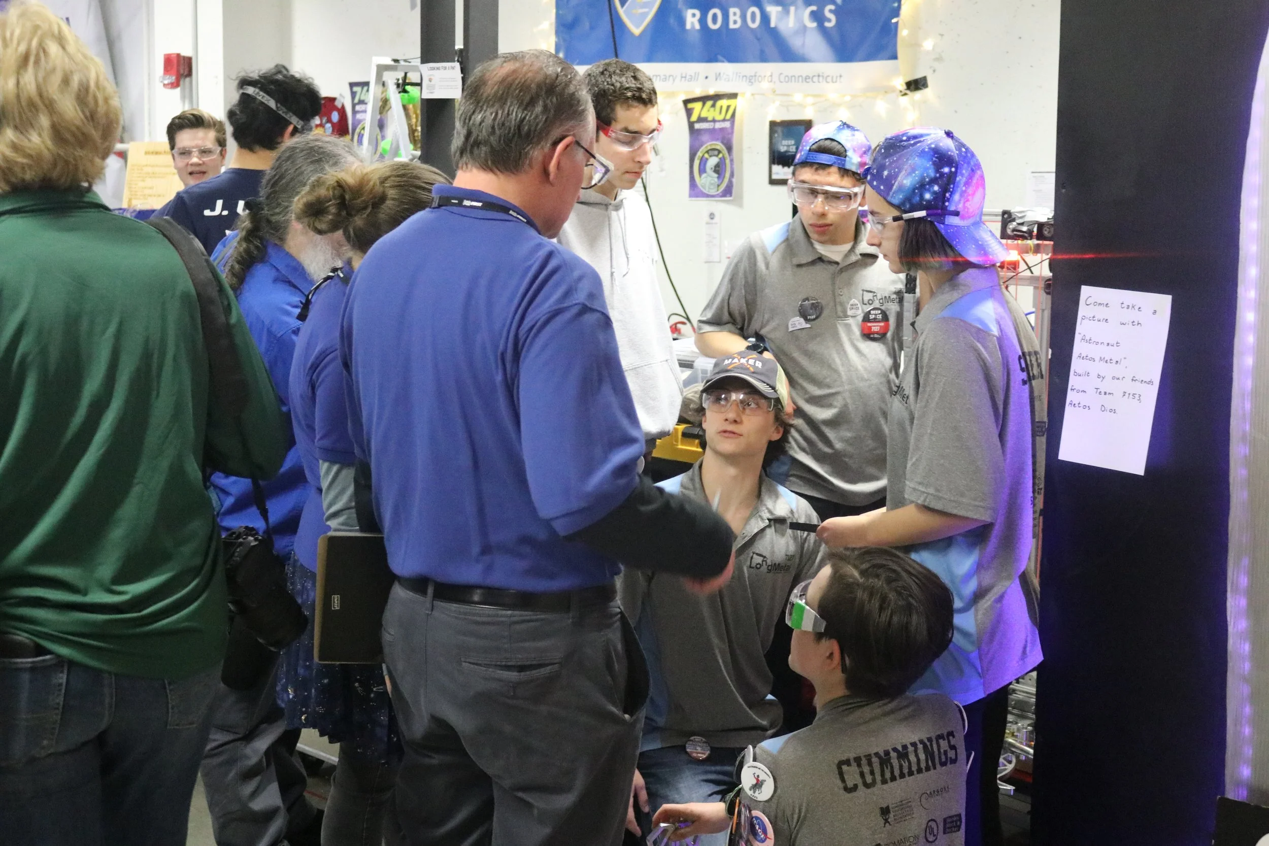 Group of students and an adult gather around a table at a robotics event. Some students are seated, wearing safety goggles and team shirts, while others stand, discussing a project. A banner with the word 'ROBOTICS' is visible above them.