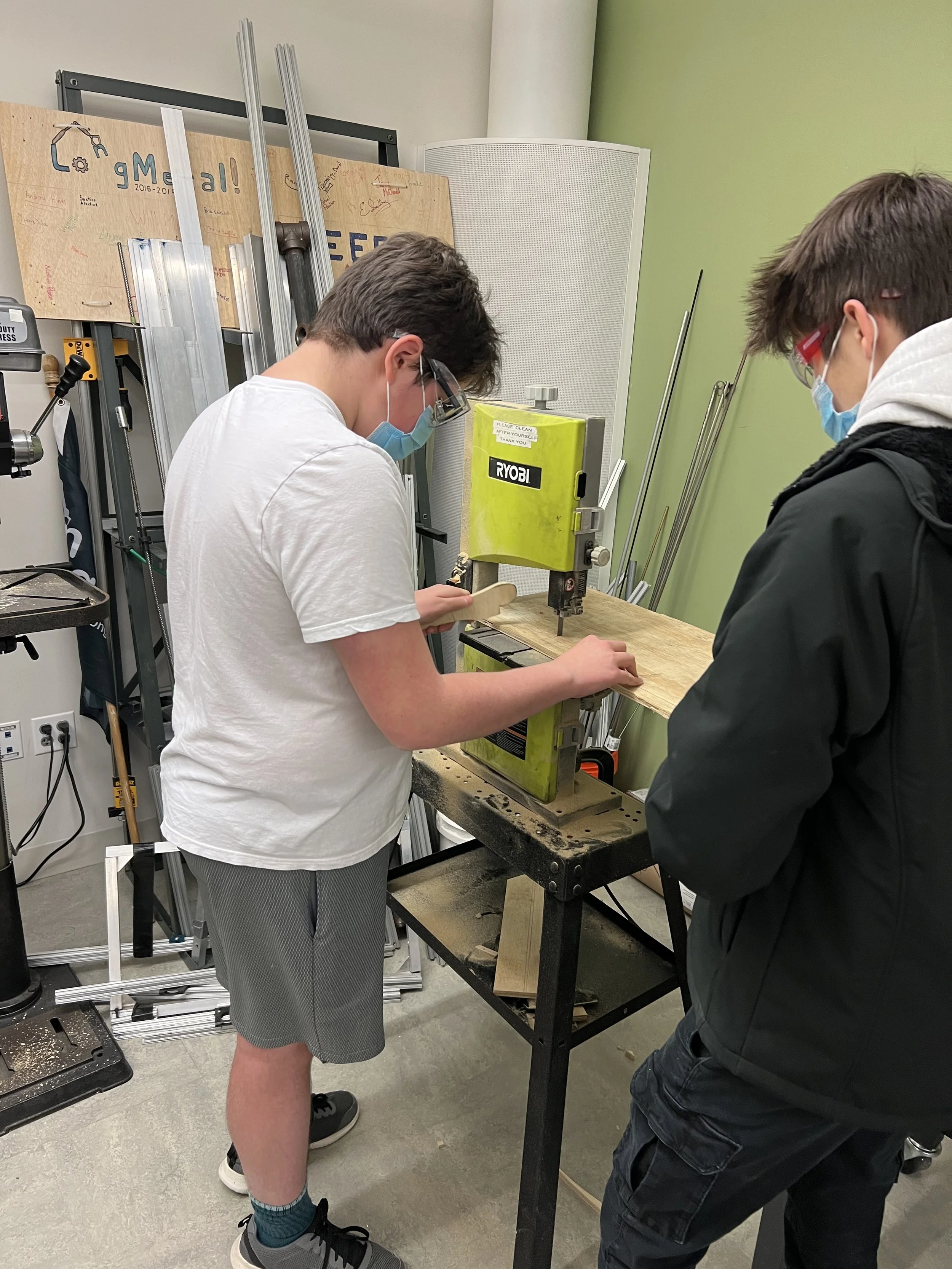 Two young boys wearing safety glasses and face masks working on a woodworking project using a Ryobi drill press at a school woodworking class.