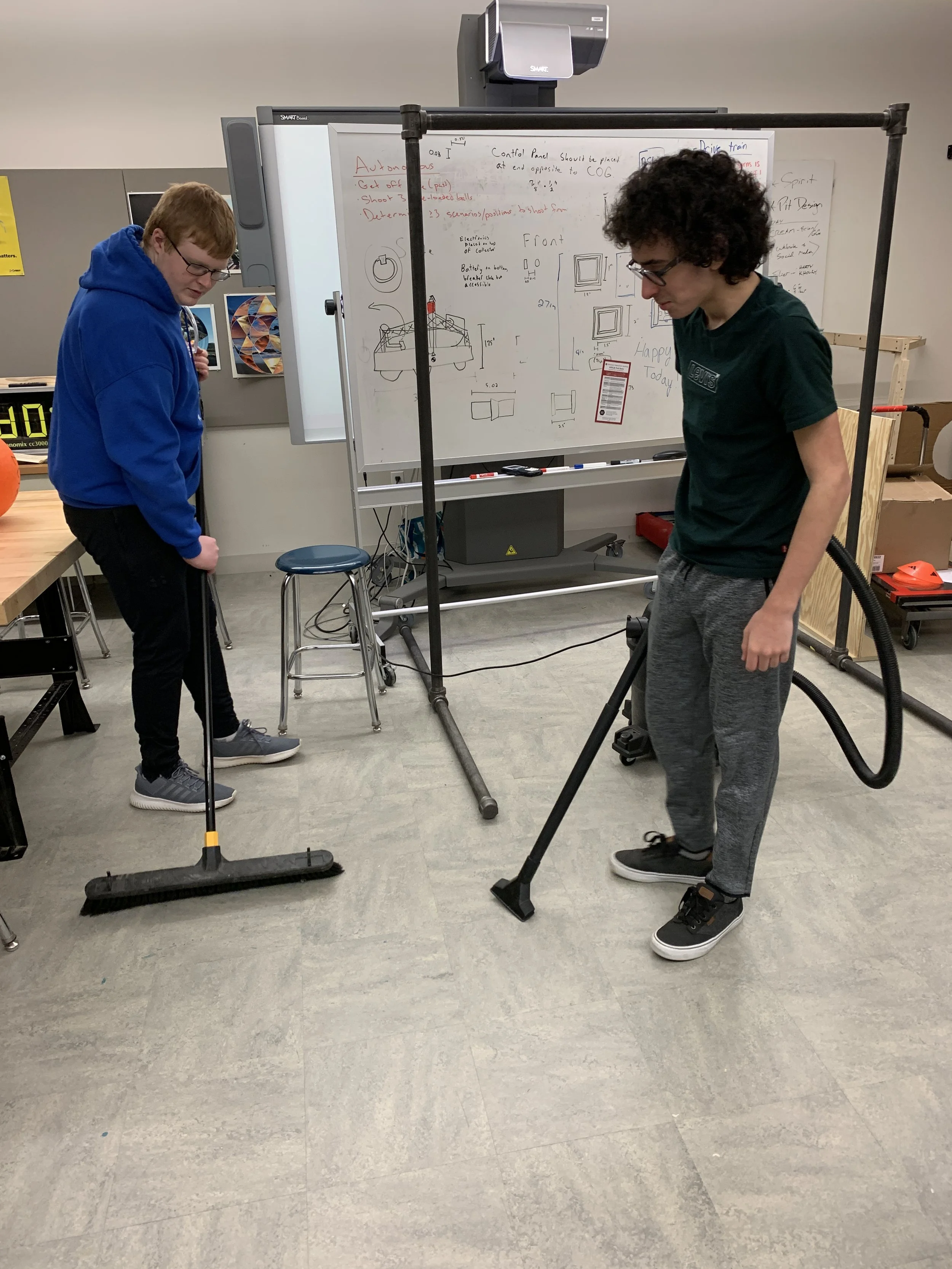 Two young men working on a project, one sweeping the floor and the other vacuuming, with a whiteboard containing sketches and notes behind them.