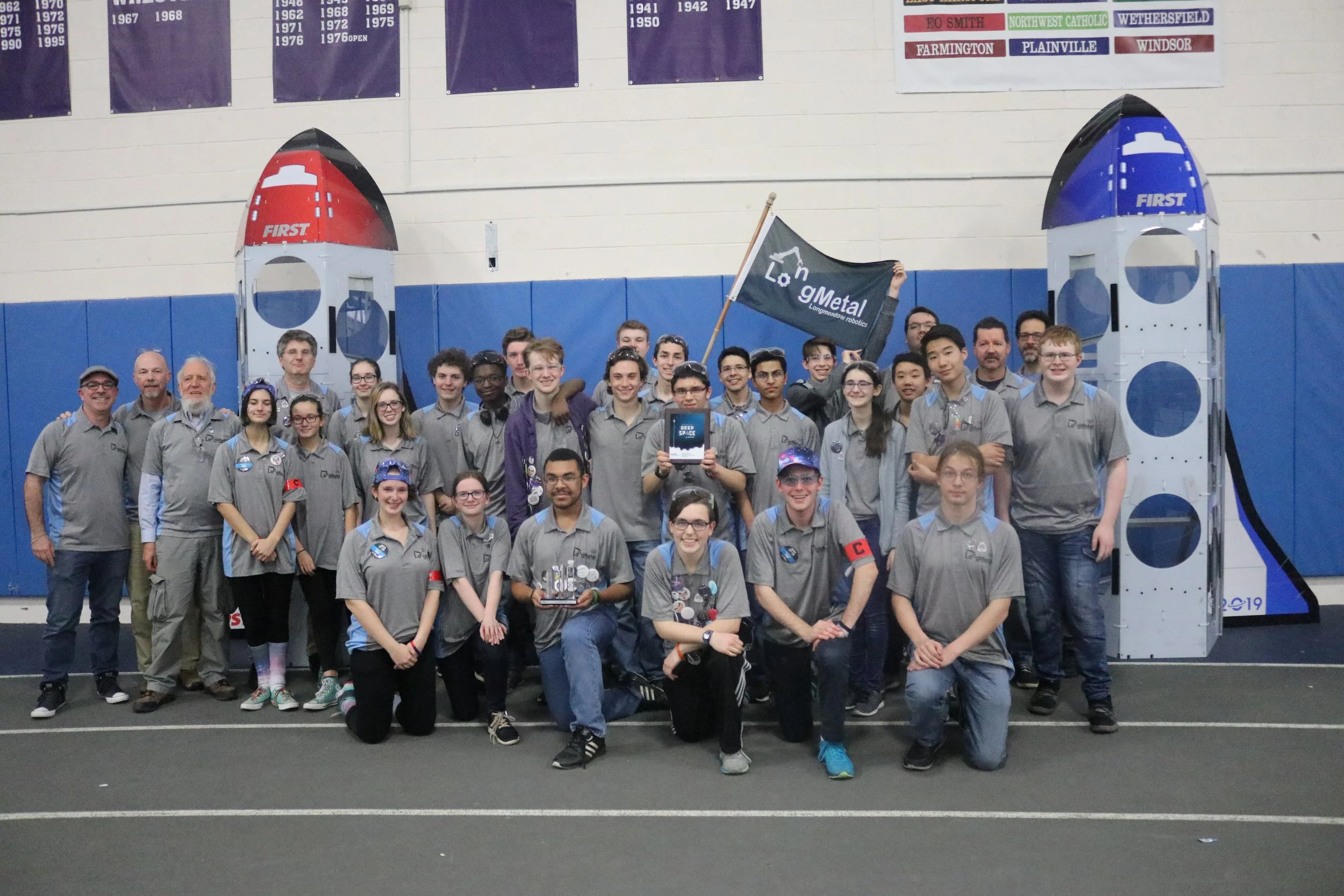 Group of students and adults posing at a robotics competition, with two rocket-shaped structures behind them. Some individuals are holding awards, and a person at the back is holding a flag with a gear logo and the words 'LongMetal'.
