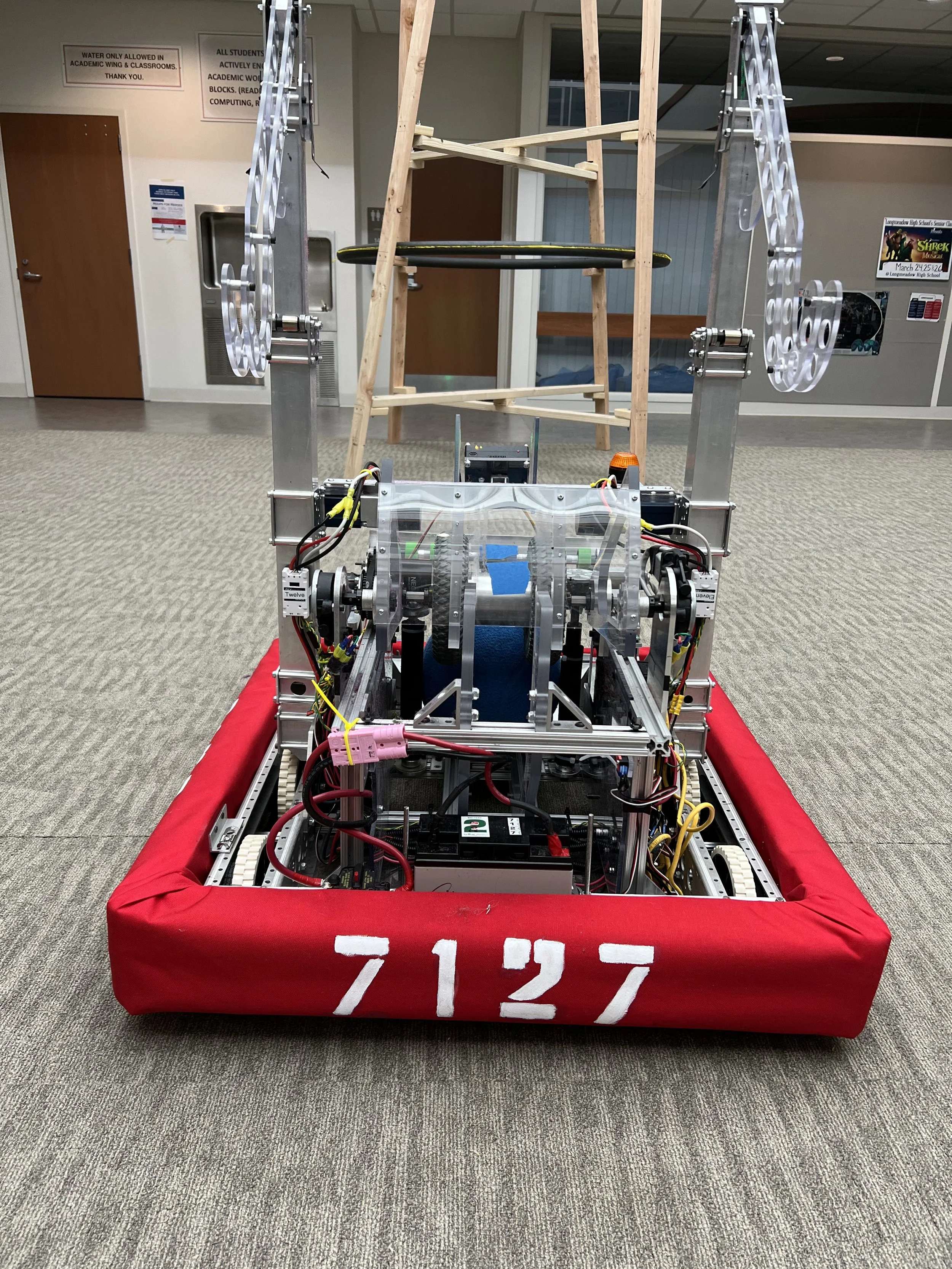 A robot with mechanical arms and a frame, labeled with the number 7127, is positioned on a carpeted floor in an indoor setting, possibly a school or laboratory. The robot has wires and electronic components visible and is surrounded by a red bumper.