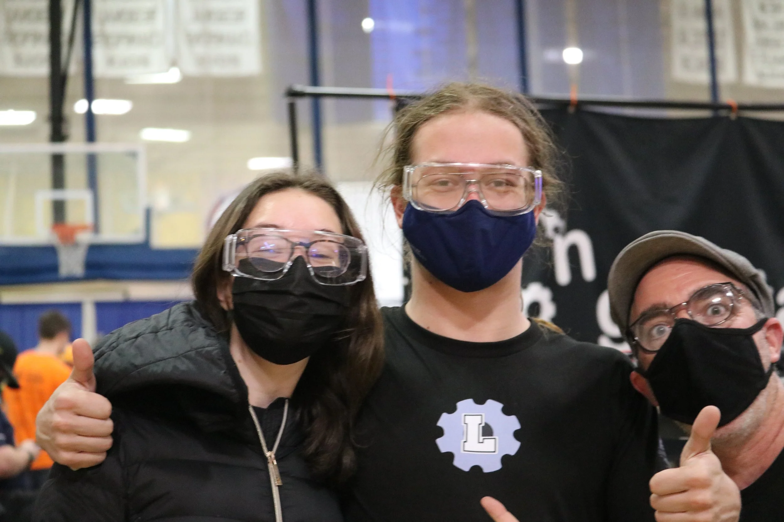 Three people wearing masks and safety glasses, giving thumbs up at a sports event in a gymnasium.
