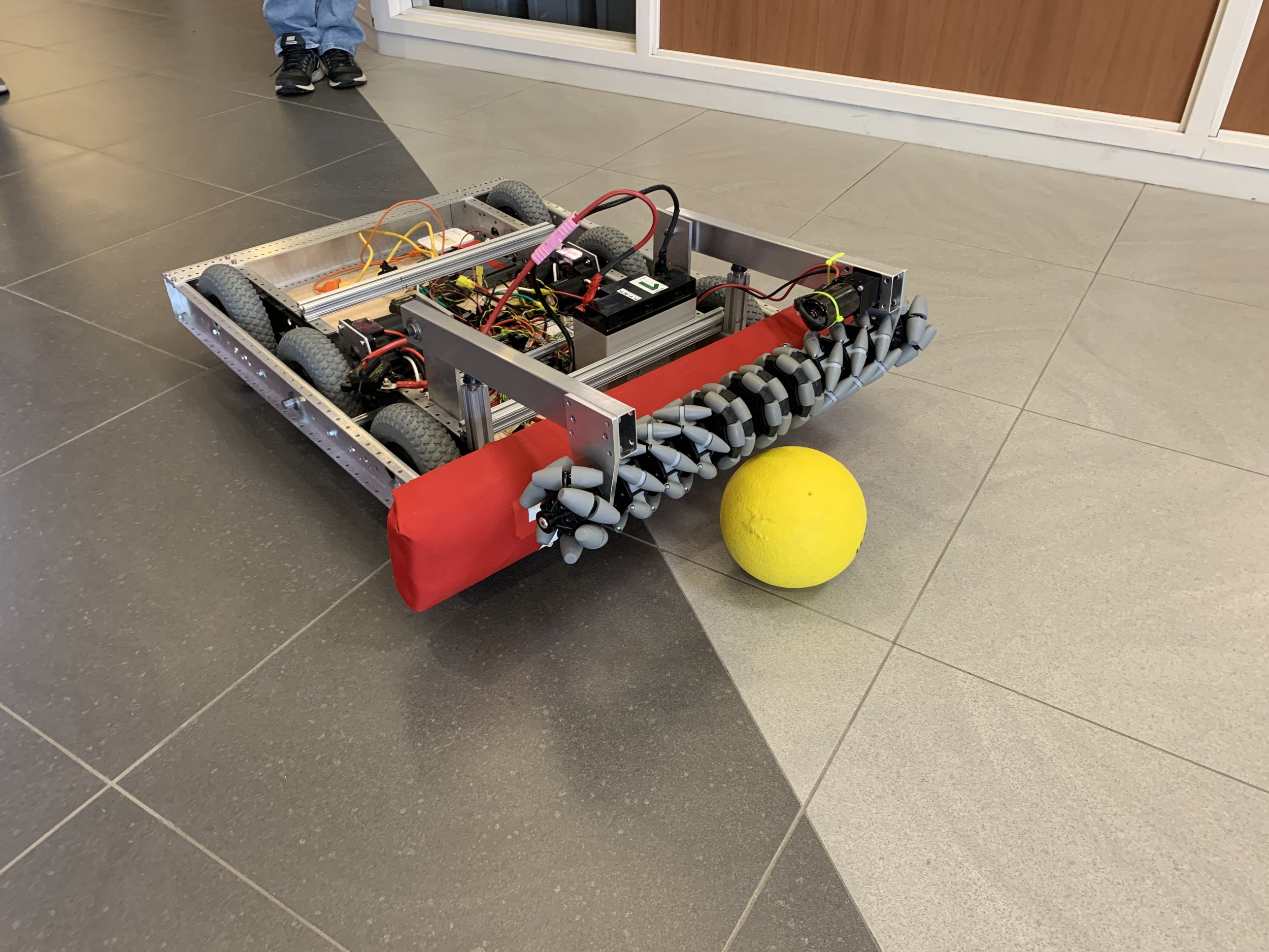 A robotic vehicle with wheels and electronic components, equipped with a red bumper, chasing a yellow ball on a tiled floor.