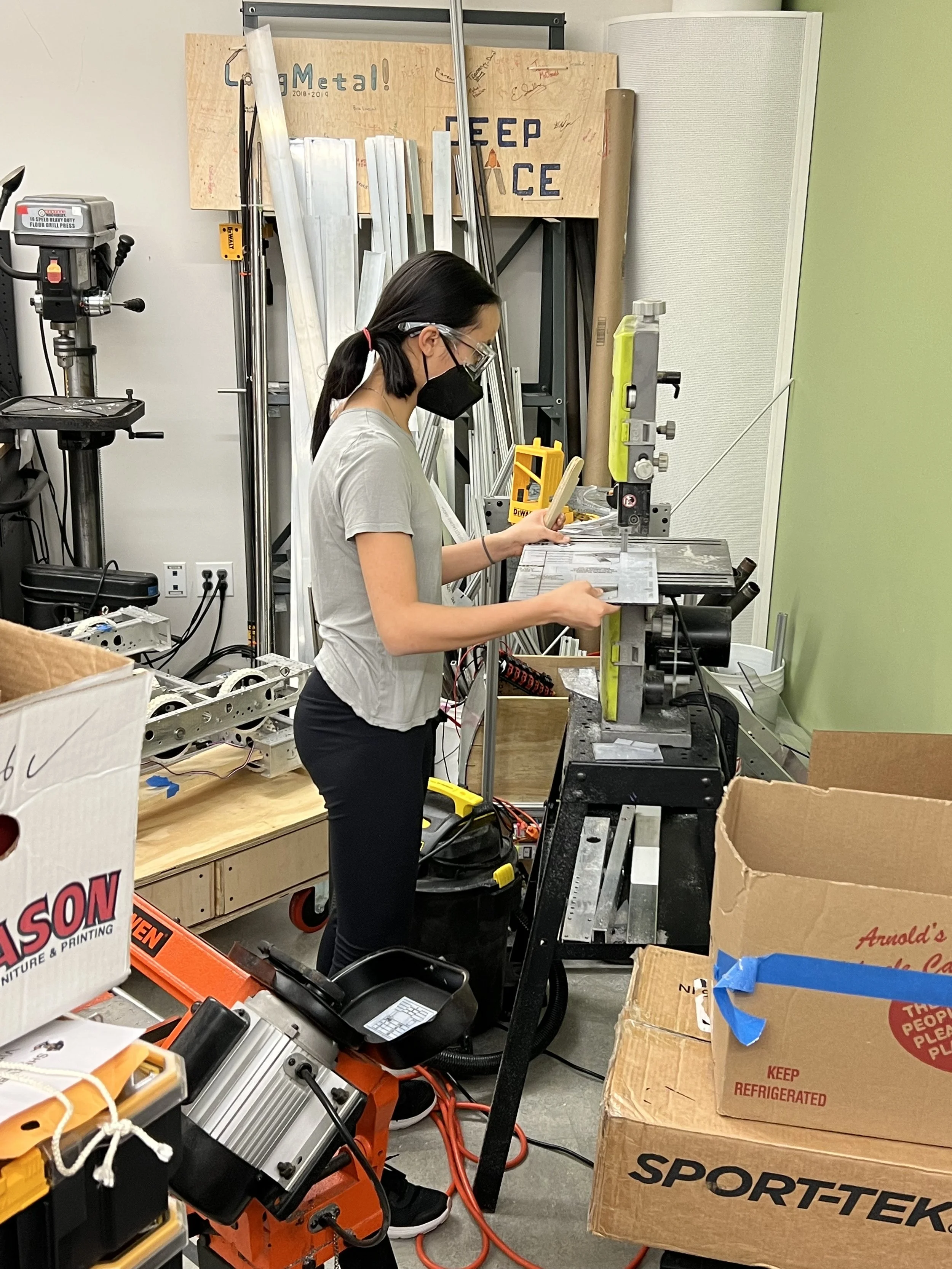 A woman wearing a black face mask, glasses, and casual gray shirt operating a metalworking machine in a workshop filled with tools, equipment, and cardboard boxes.