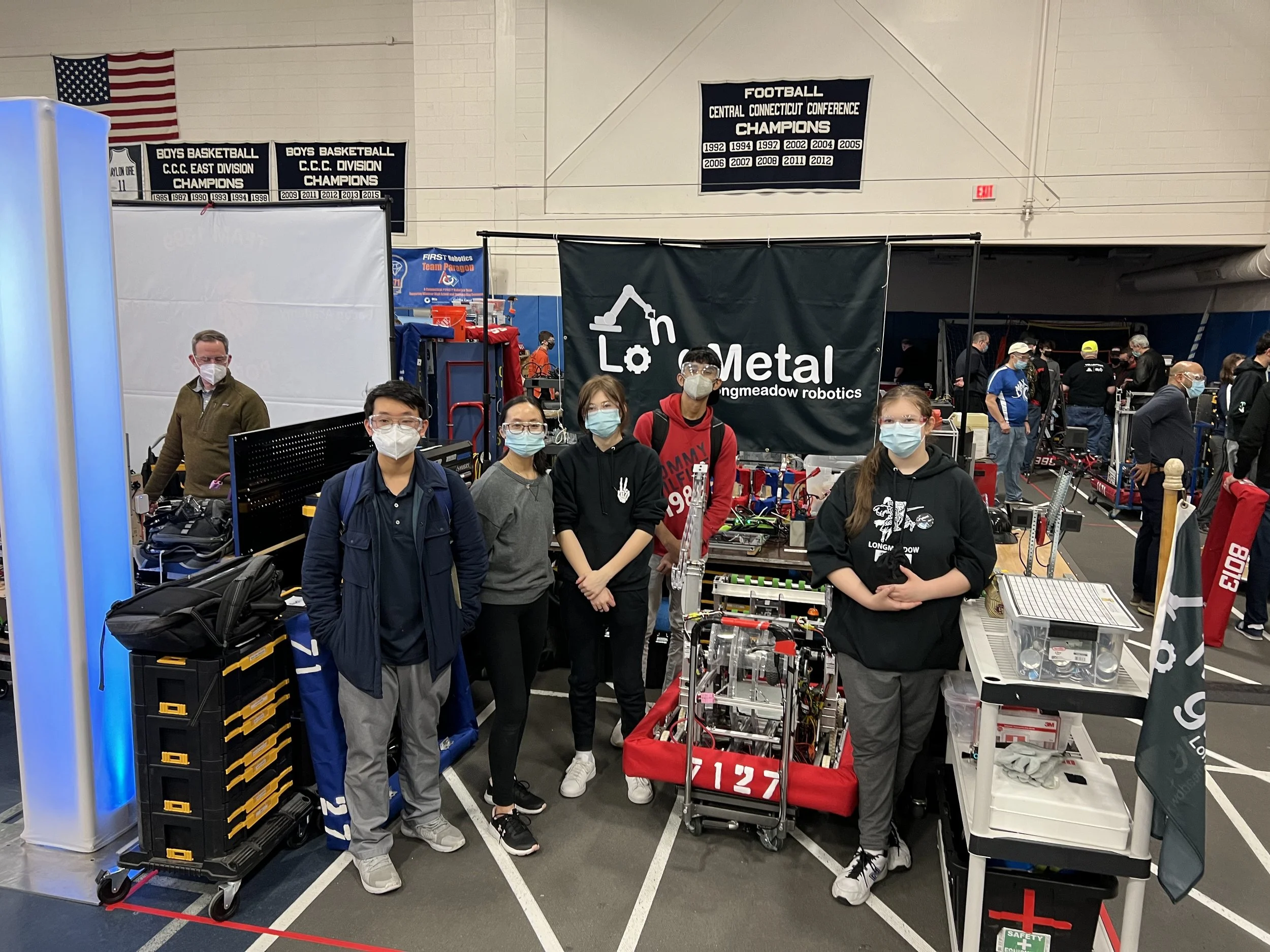 Group of five young people wearing masks and casual clothes, standing in front of a robotics display at a competition, with a banner that reads Longmeadow robotics, and various robotic equipment around them.