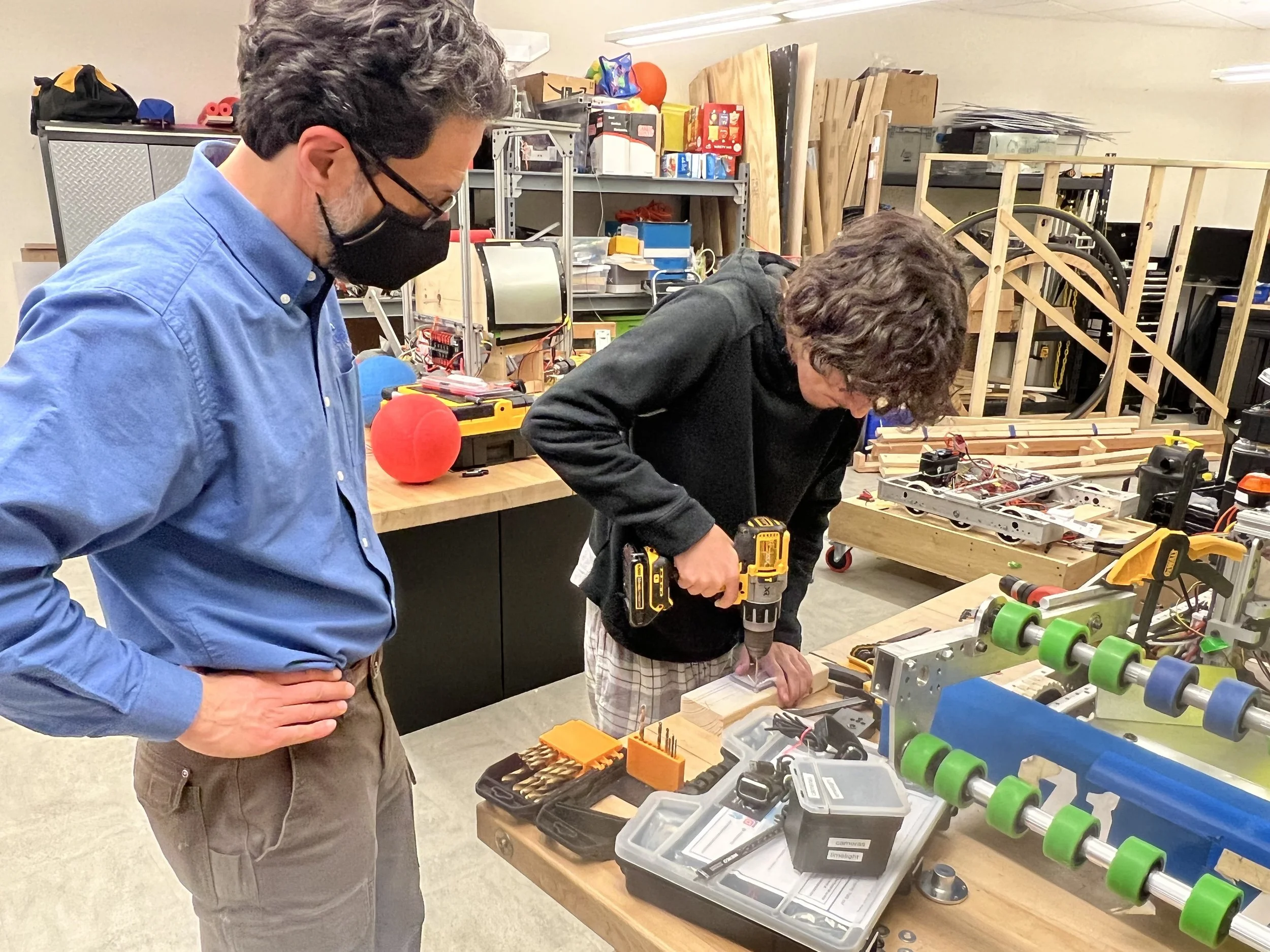 Two men working on a woodworking project in a workshop, using a power drill on a piece of wood.