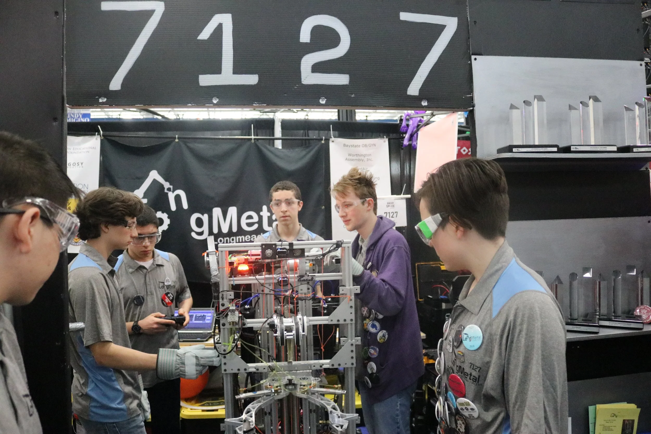 Group of students working on a robotics project at a competition, with a black banner behind them displaying the word 'Longmetal' and hardware on shelves around them.