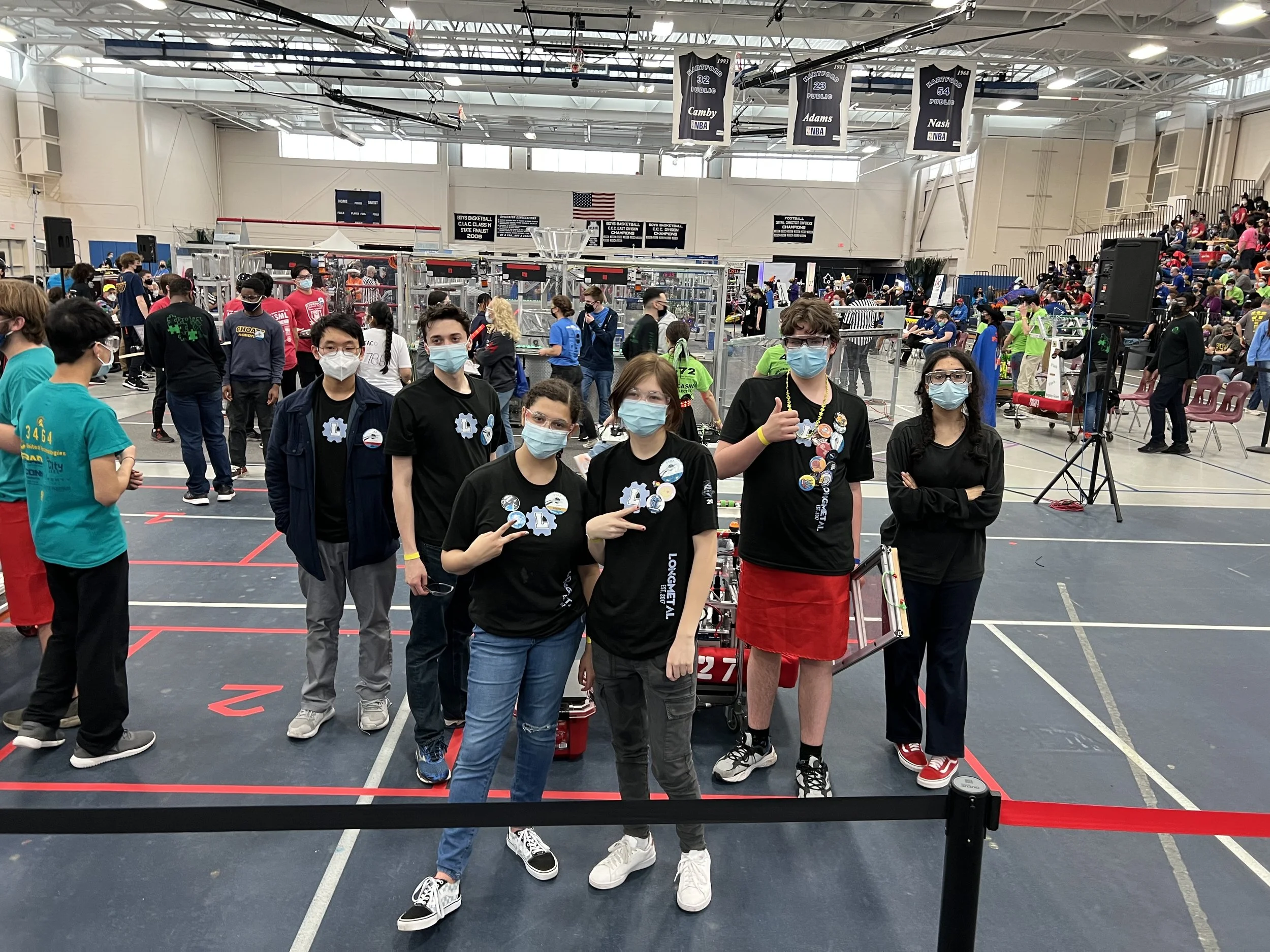 Group of six students and a teacher in a gymnasium at a robotics competition, wearing masks and posing in front of robot and competition area.