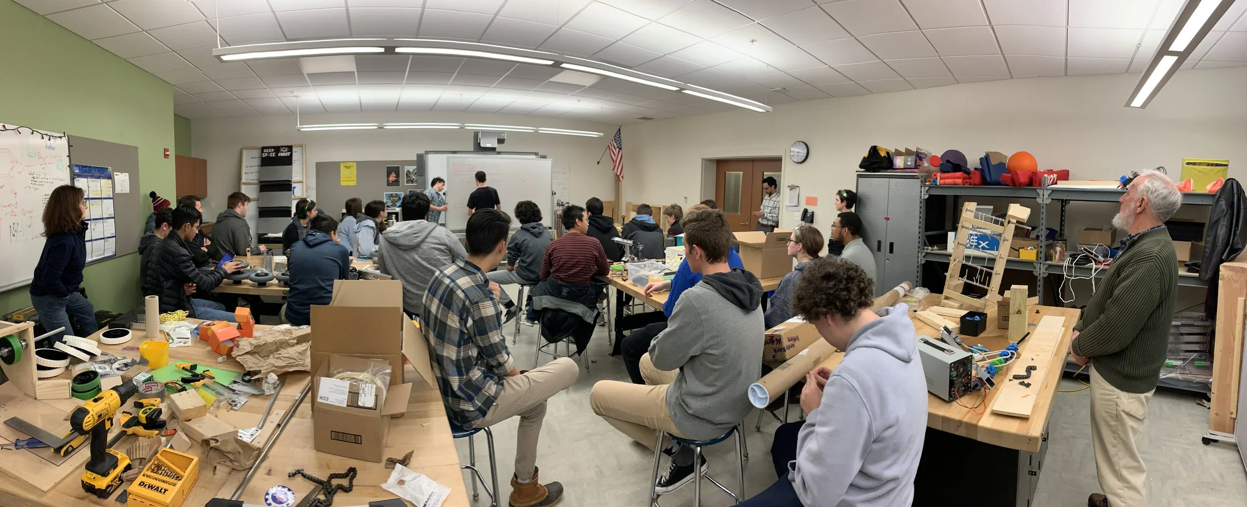 A classroom filled with students and instructor during a workshop, with tables, tools, and supplies, and a whiteboard at the front.