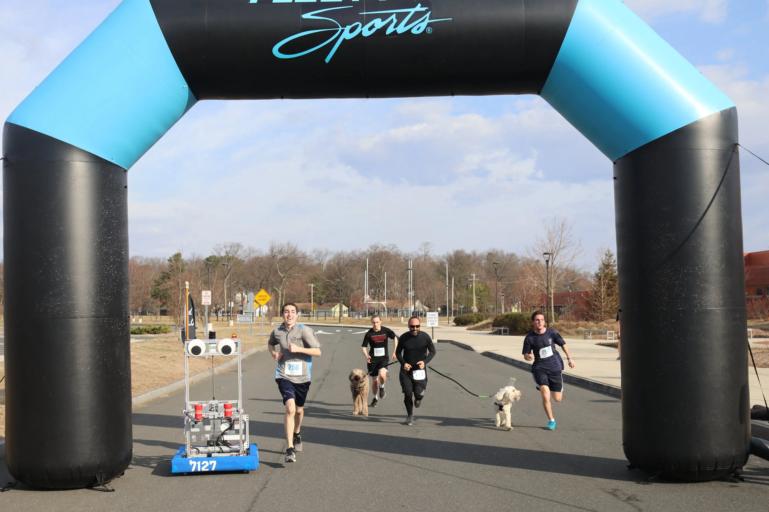 People running in a race under an inflatable archway with the word 'Sports' written on it. The runners are accompanied by dogs, and there is a robot near the starting point.