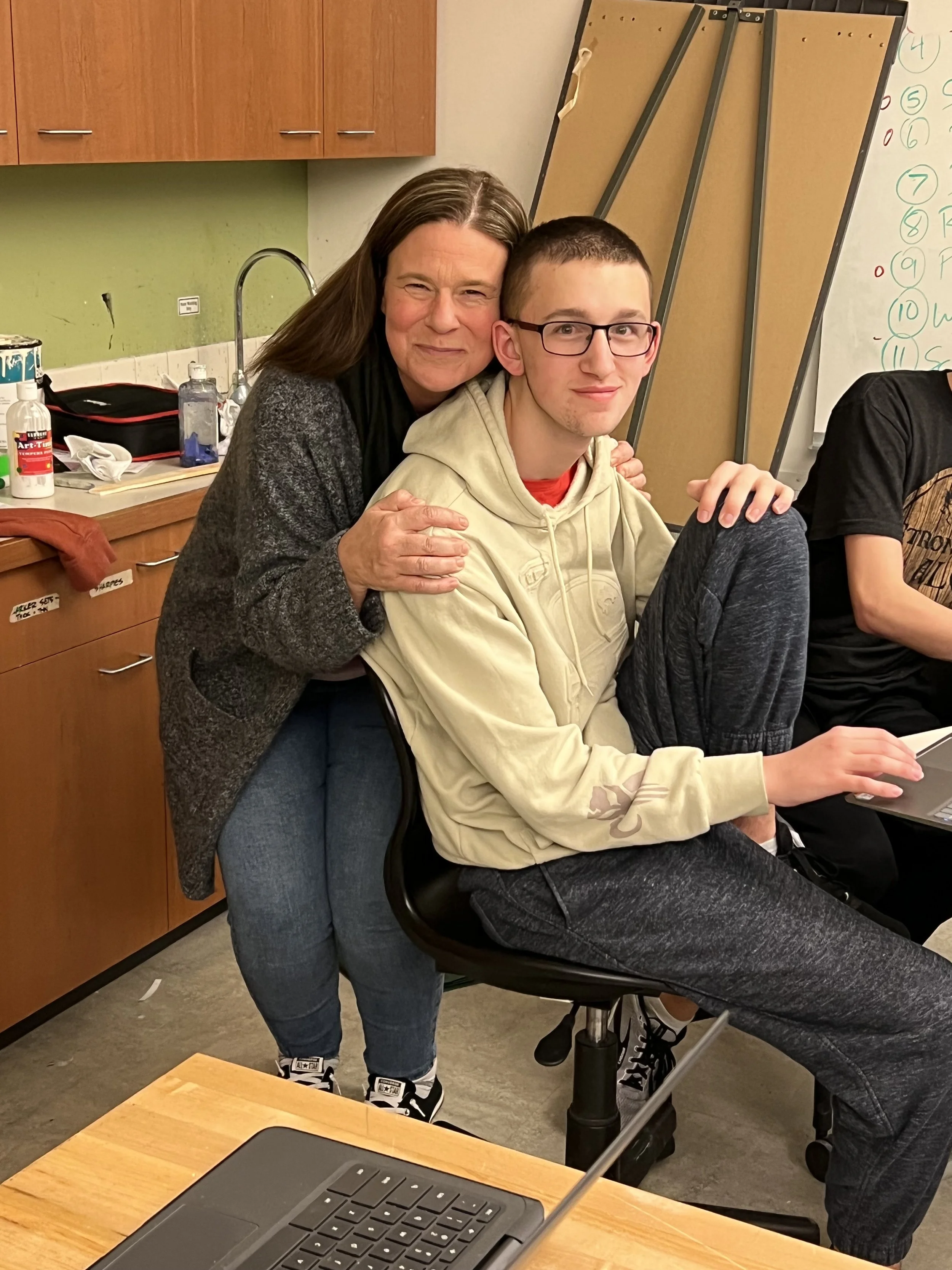 An adult woman hugging a teenage boy sitting on a chair in a classroom setting.
