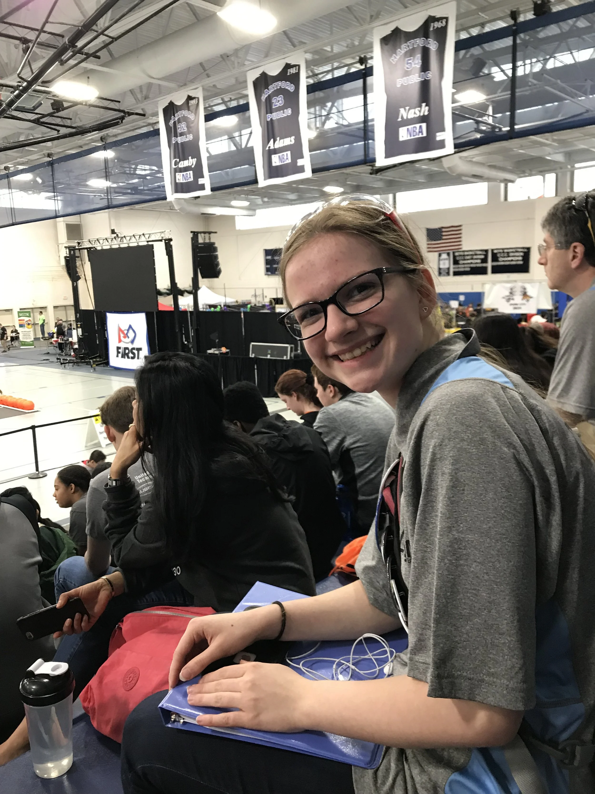 Smiling teenage girl with glasses sitting in bleachers at a robotics competition, holding a notebook and headphones, with banners hanging from the ceiling and a stage in the background.