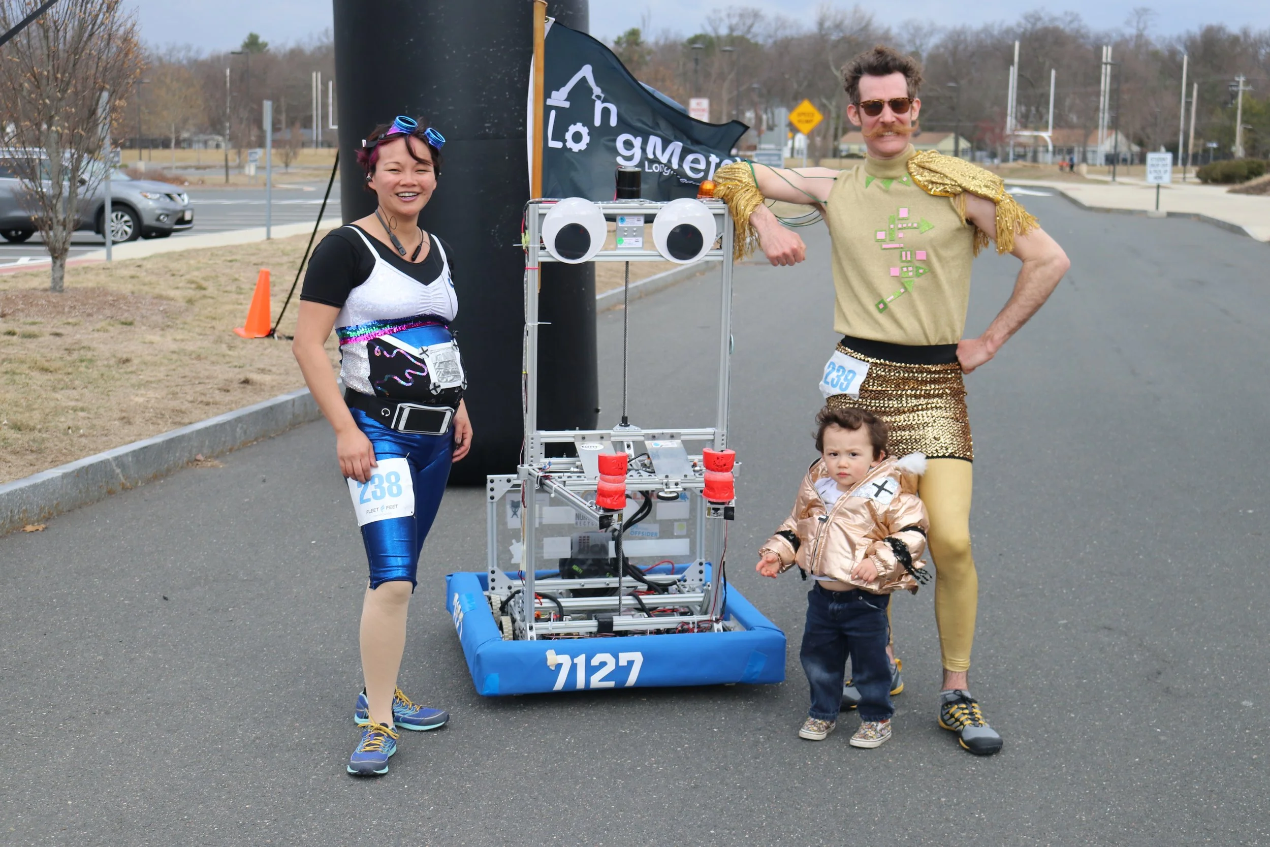 Group of four people, two adults, one child, and one robot, posing outdoors during a race or event. The adults are dressed in colorful, eccentric costumes, and the child is wearing a beige jacket. They are standing next to a robot with a blue contain