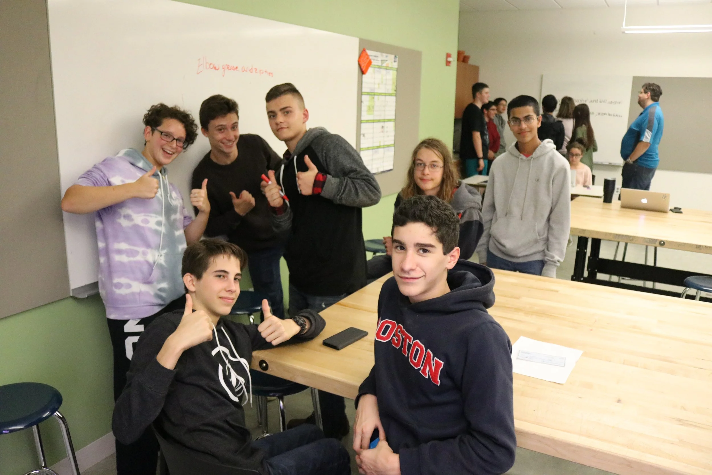Group of seven teenagers gathered around a table in a classroom, some sitting, some standing, smiling and showing thumbs up; background includes other students and a whiteboard.
