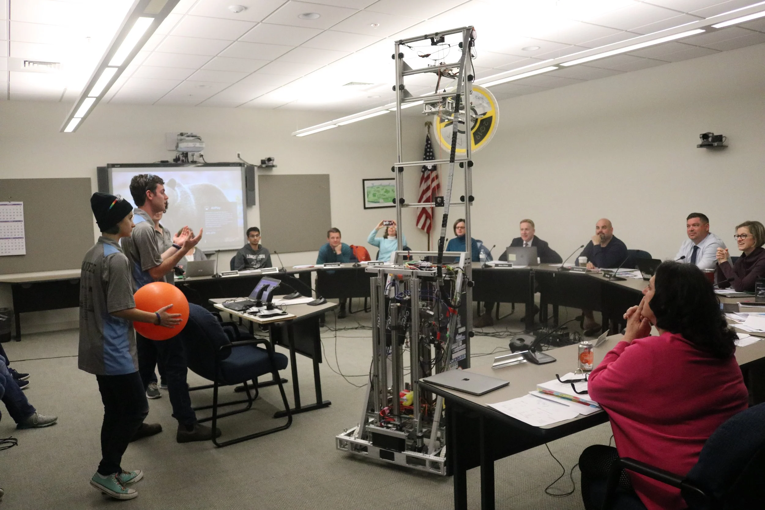 Students presenting a robotics project to a panel in a conference room, with a large robot structure in the center.