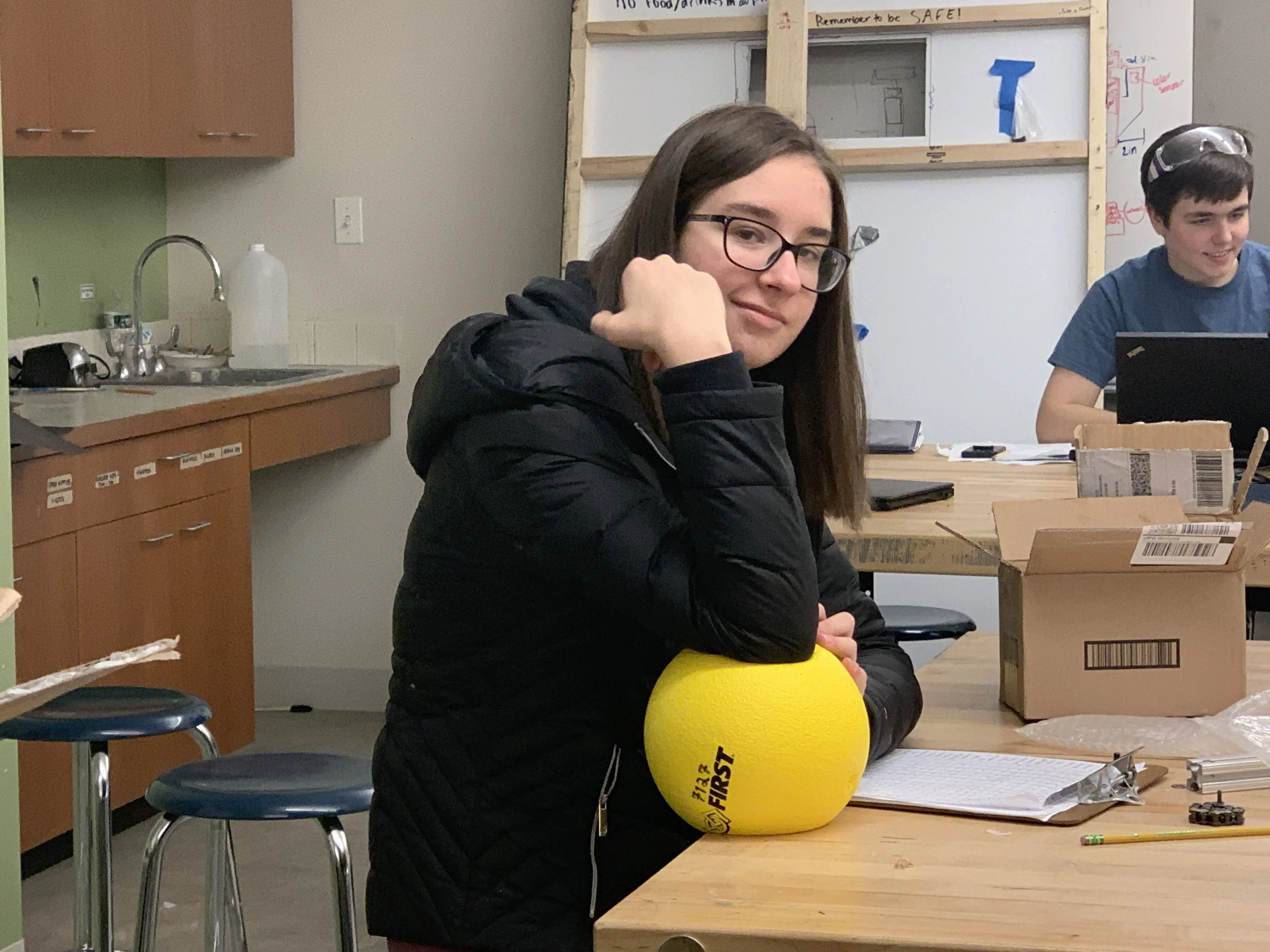 A young woman with glasses and long brown hair sits at a wooden table in a classroom or lab, resting her head on her hand, with a large yellow foam ball labeled 'FIRST' in front of her. She is wearing a black jacket. Behind her, a boy in a blue shirt