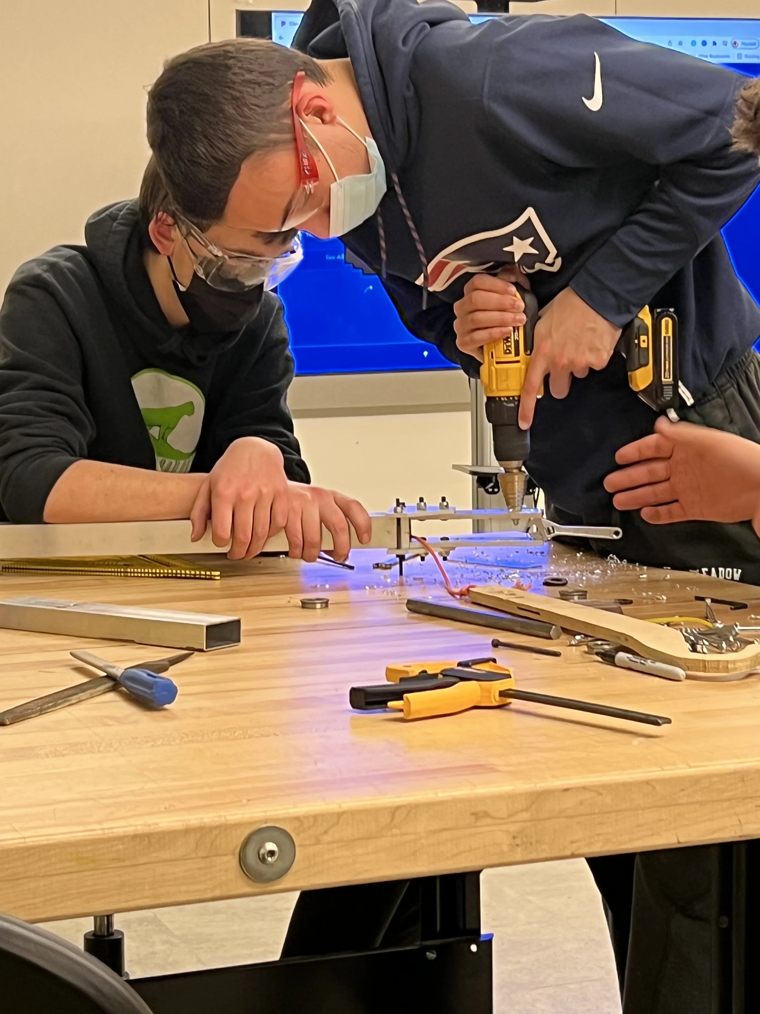 Two young men working on a small project at a wooden table, one using a power drill. Both are wearing masks and safety glasses, with various tools and materials on the table.