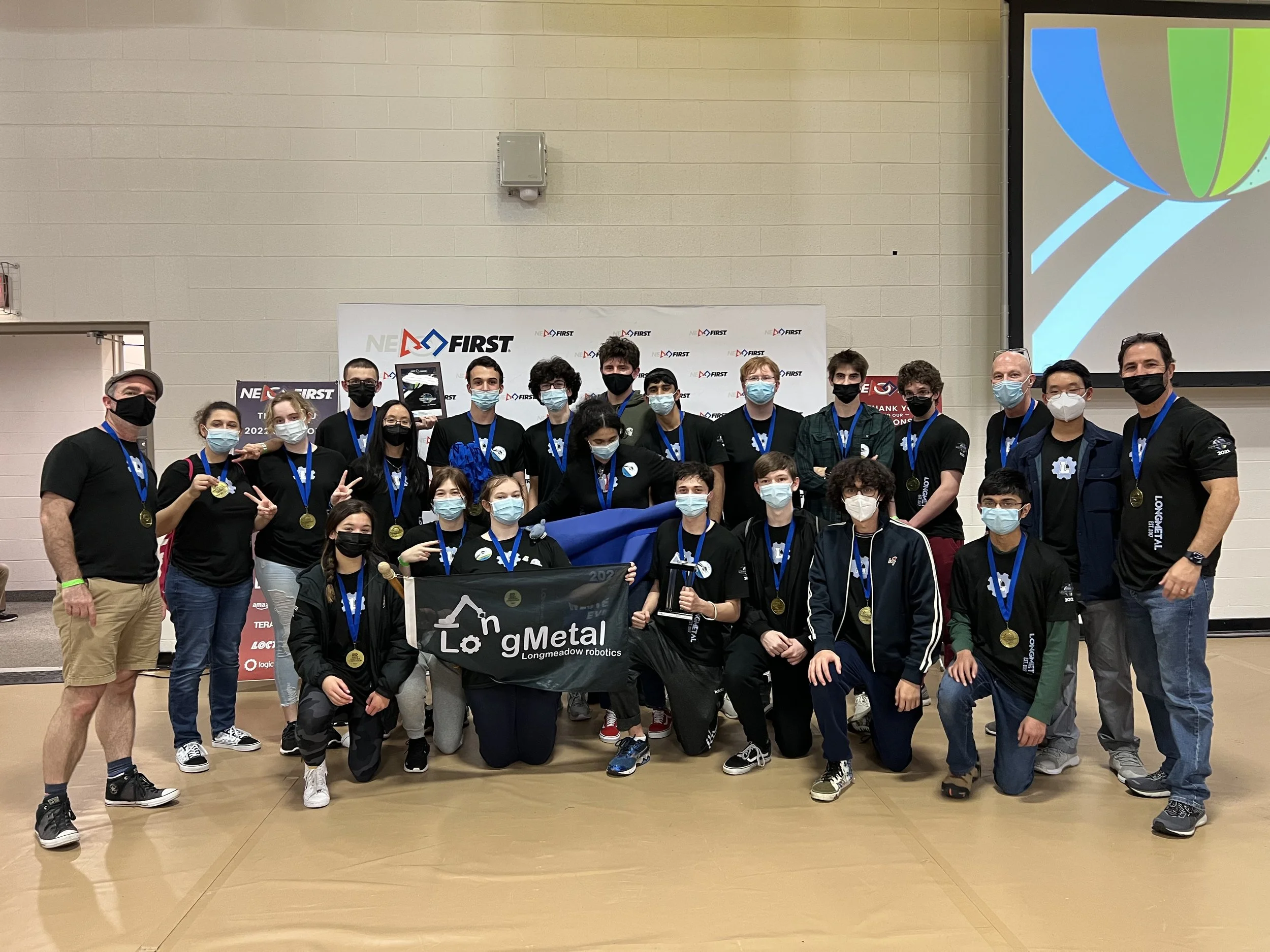 Group of young individuals and coaches posing with medals at a robotics competition, holding a banner that reads "Longmeadow robotics." They are wearing face masks and standing in front of a backdrop with the FIRST logo and a large screen displaying 