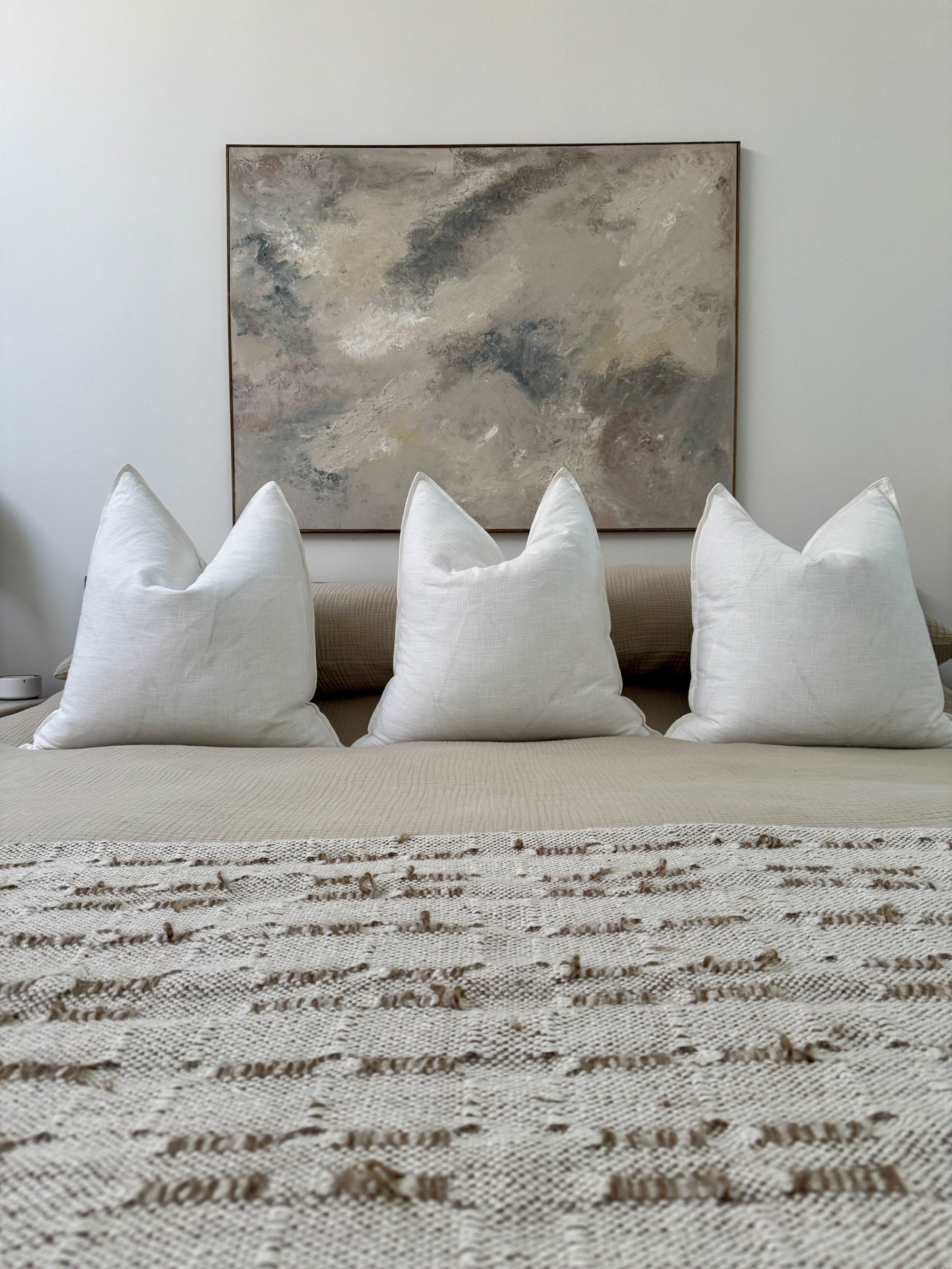 A beige sofa with three white pillows in front of a large abstract painting with gray, white, black, and beige colors on a white wall, topped with a cream and brown woven blanket.