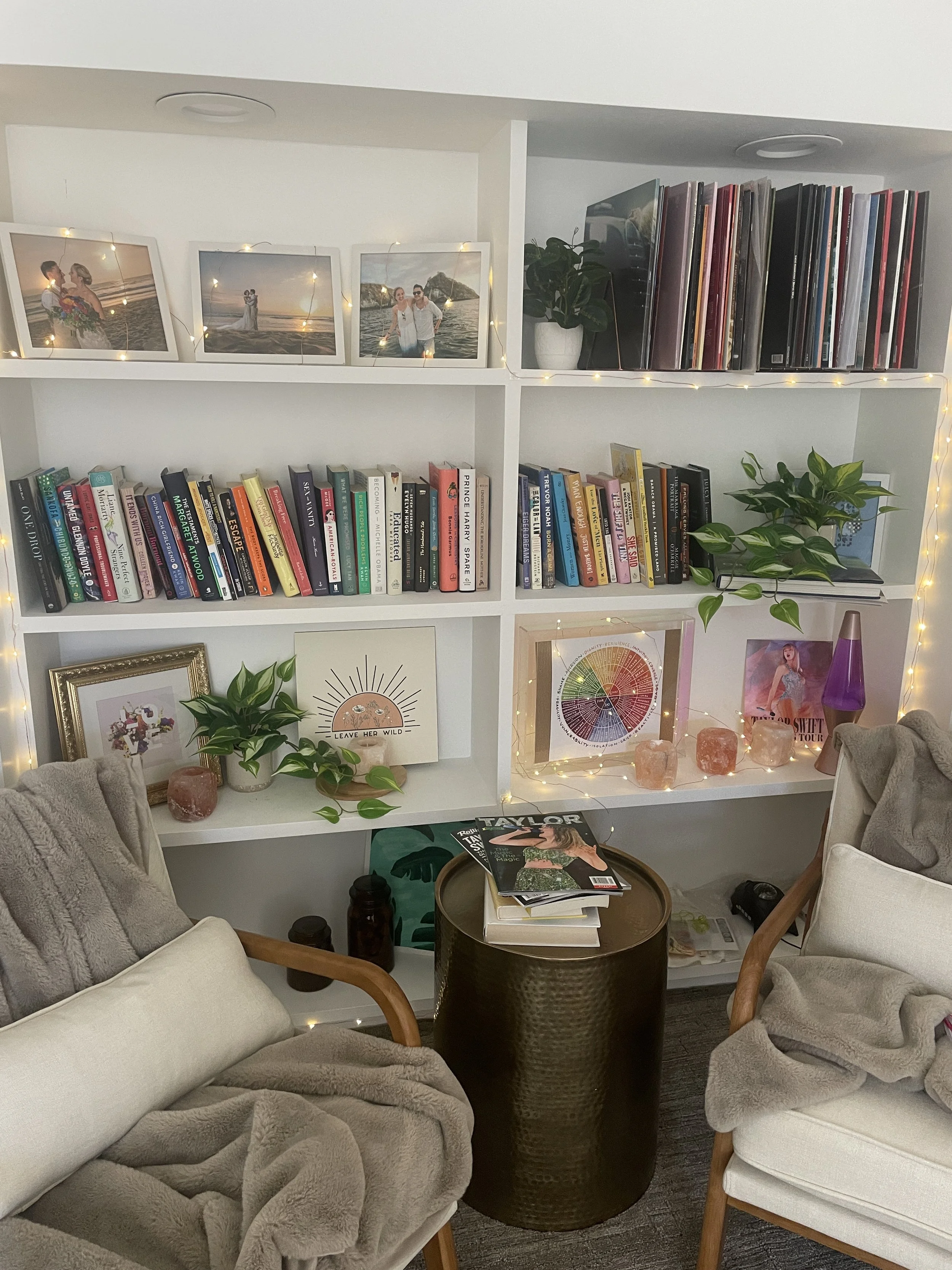 Living room scene with two beige armchairs with wooden arms and light gray blankets, a black textured round side table with magazines and decorative items, a white bookshelf filled with books, framed photos, artwork, a potted plant, and string lights