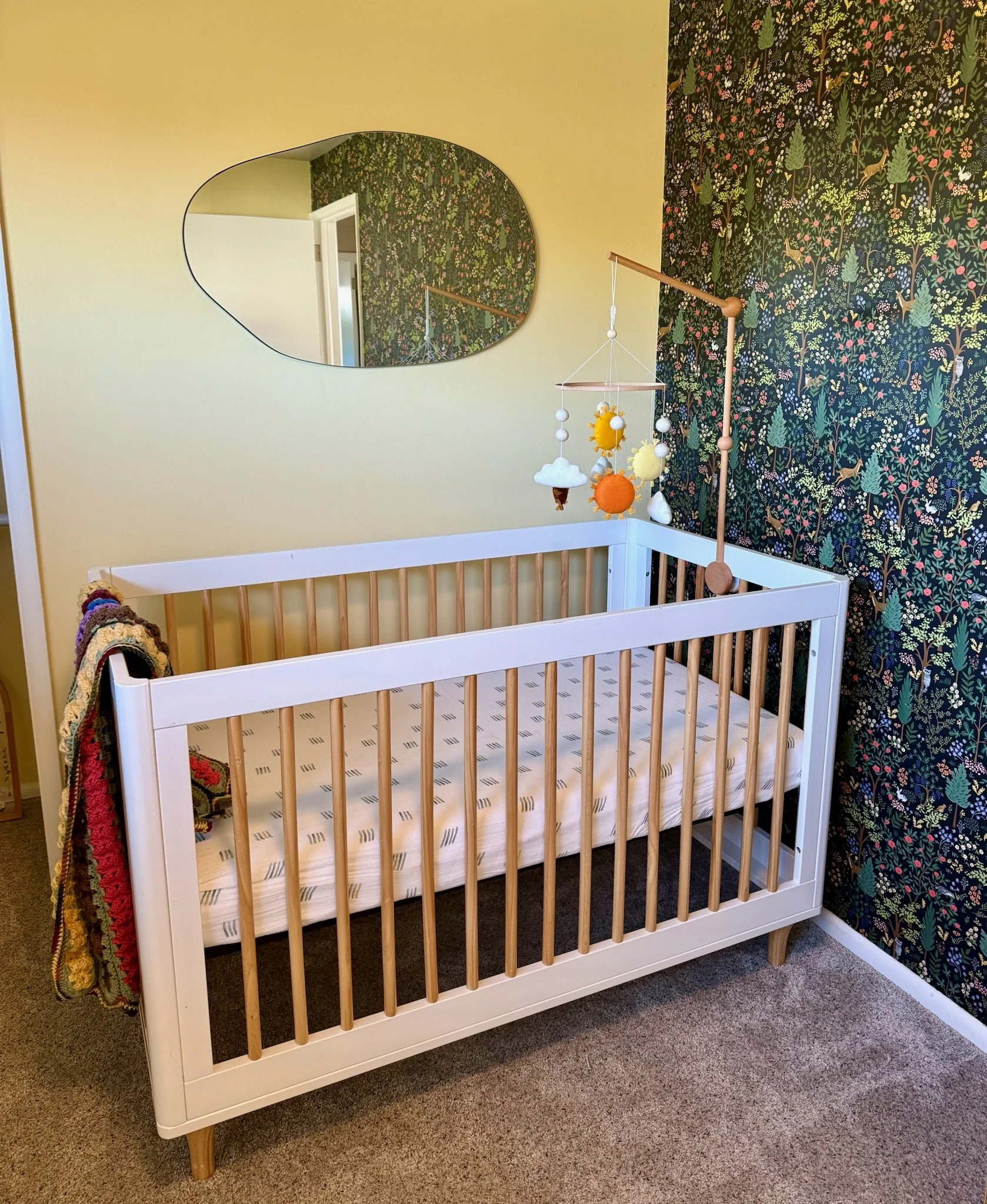 A white crib with wooden slats in a room with yellow and floral wallpapered walls, a mirror on the yellow wall, and a mobile hanging above the crib.