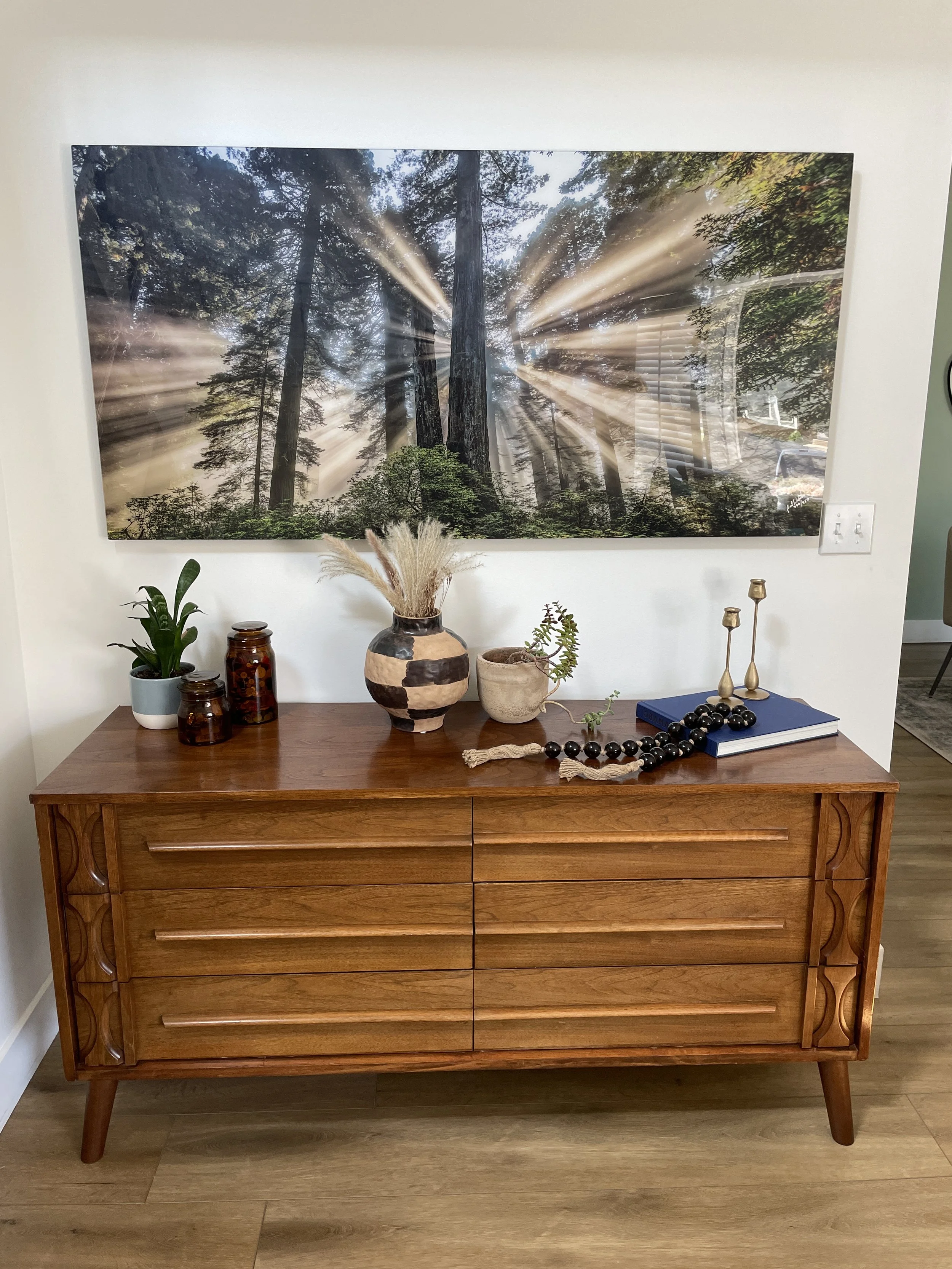 A wooden dresser with decorated plants, jars, a book, candle holders, and a large piece of art featuring trees and sun rays hanging on the wall above.