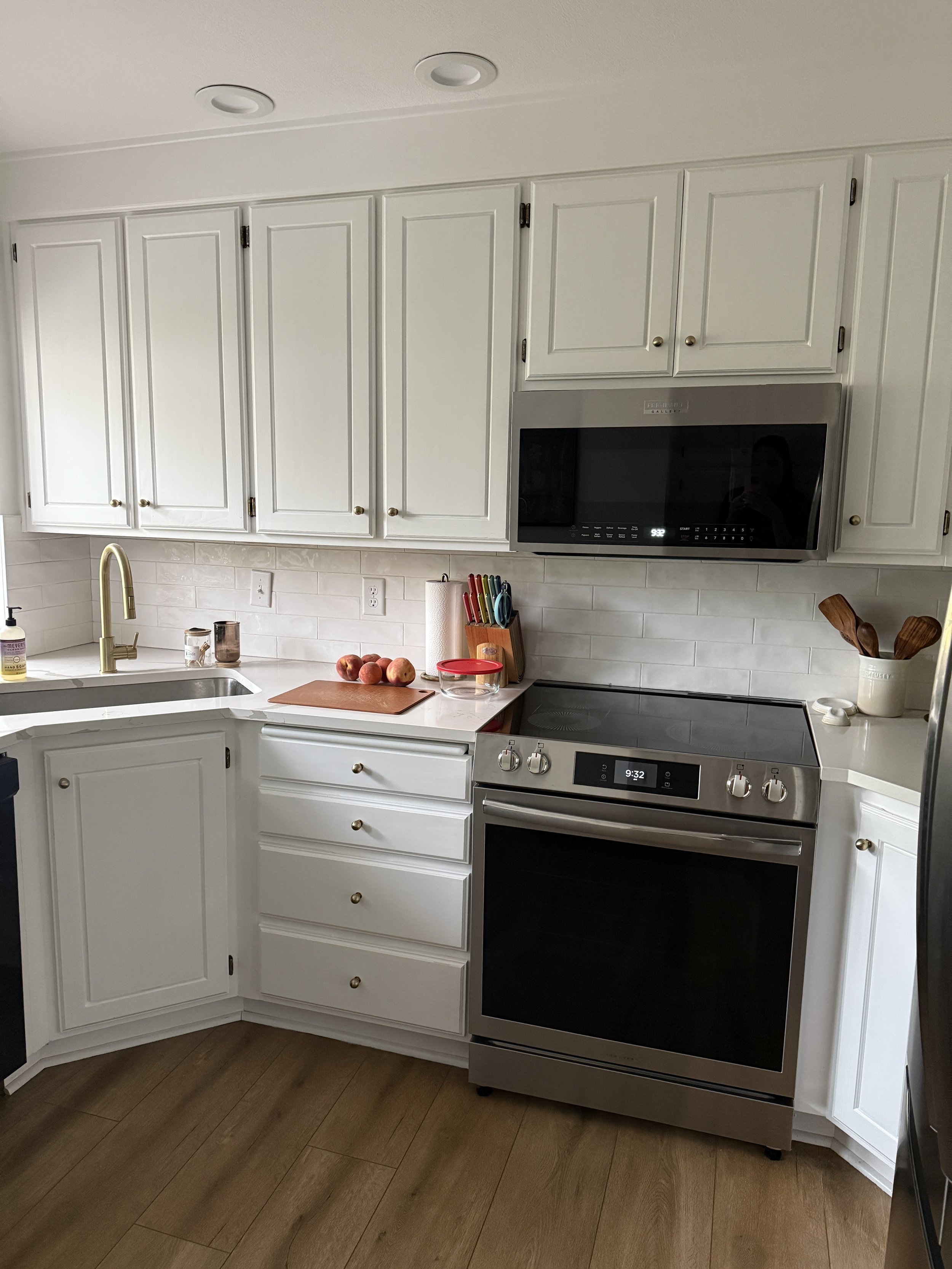 White kitchen cabinets above a countertop with a black microwave built into the cabinetry. The countertop has peaches, scissors, and a paper towel roll. A sink with a gold faucet is on the left, and a stove with digital clock is on the right. The bac