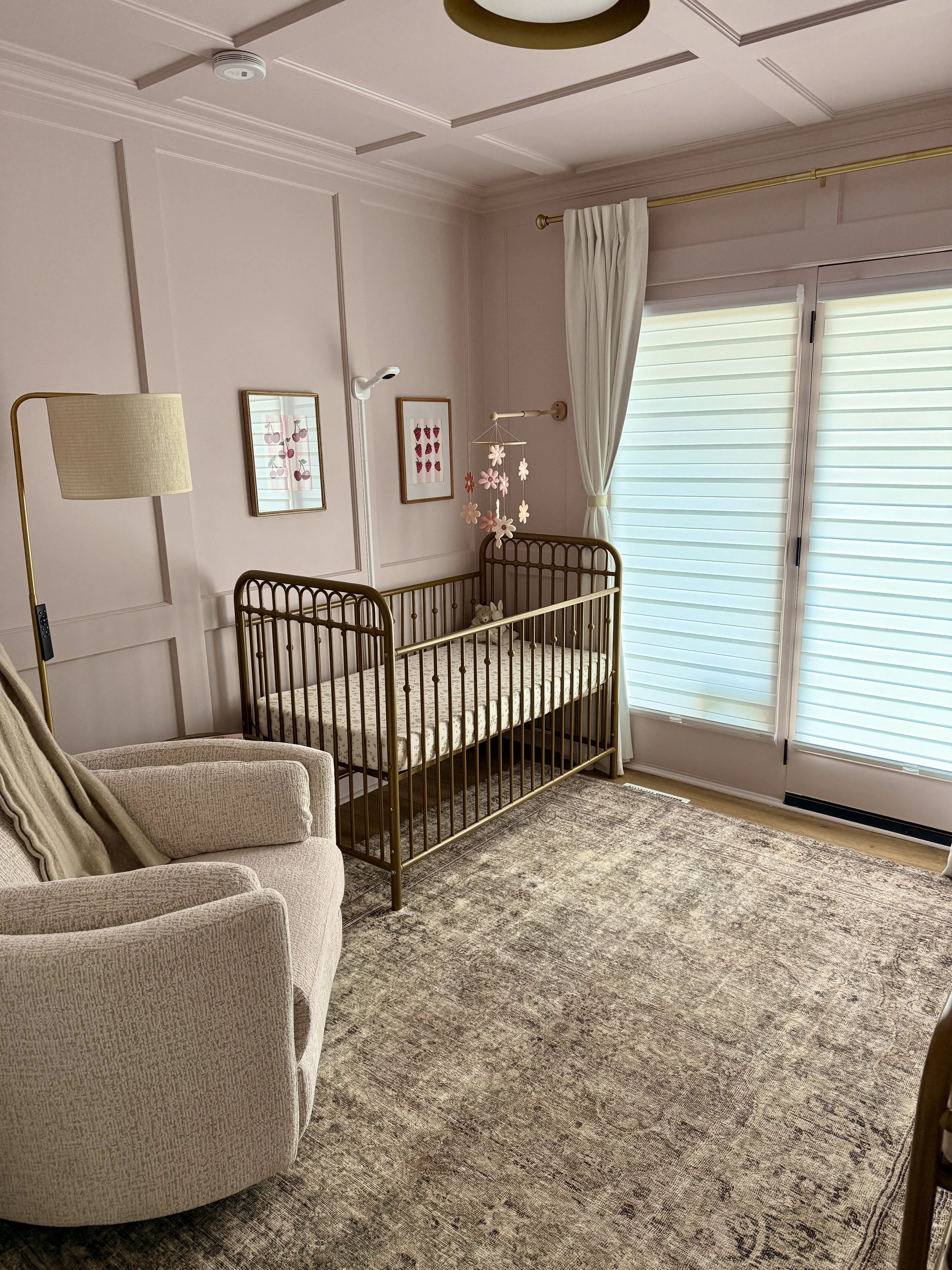 A nursery room with a beige armchair, a gold crib with stuffed animals, two framed pictures on the wall, a mobile with pink and white flowers, a window with white curtains, a wooden floor, and a large patterned rug.