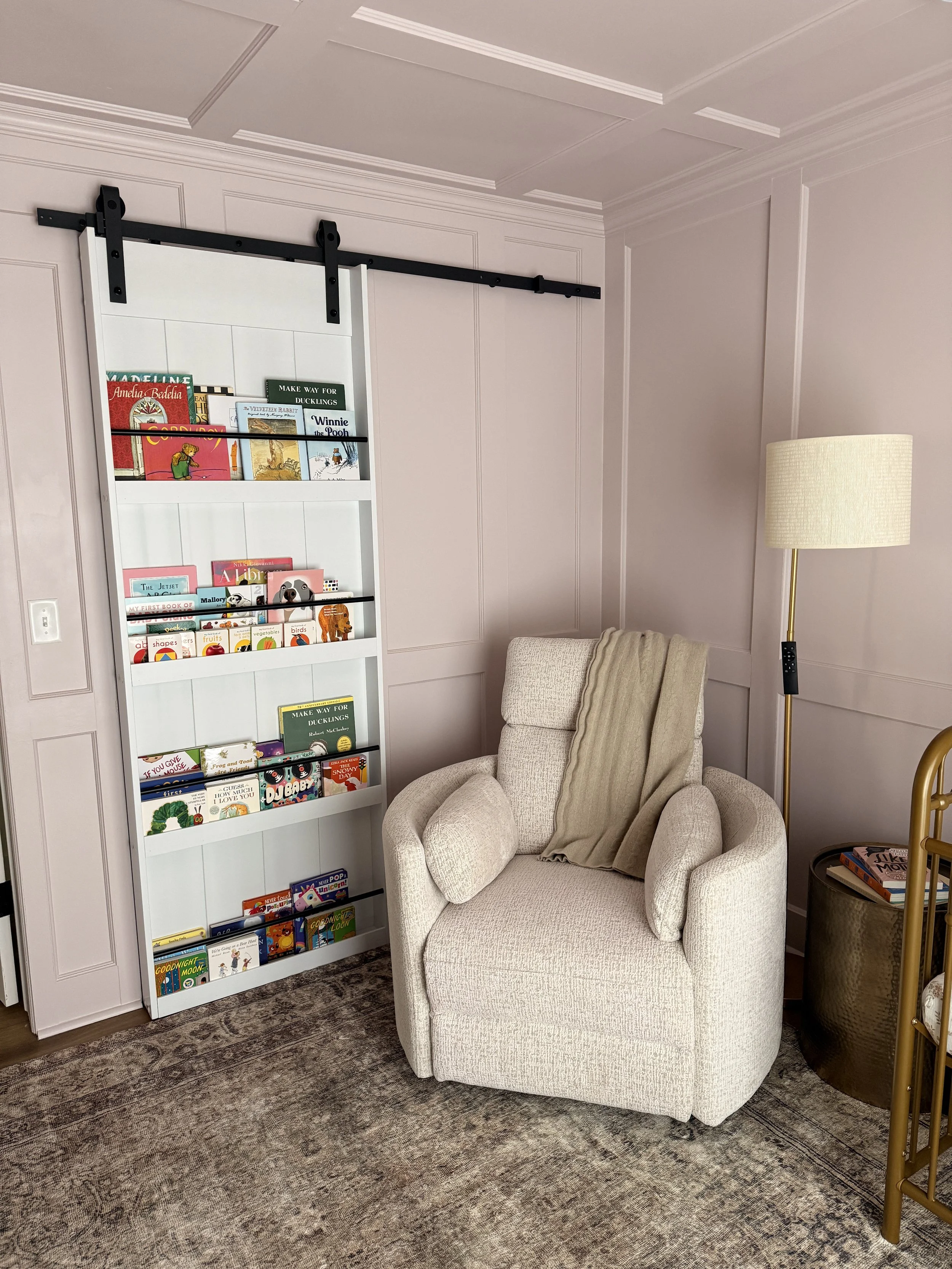 A cozy reading nook with a beige armchair and a throw blanket, a tall bookshelf filled with children's books, a standing lamp with a beige shade, and a small round side table with books on it, located in a room with light-colored walls and wood floor