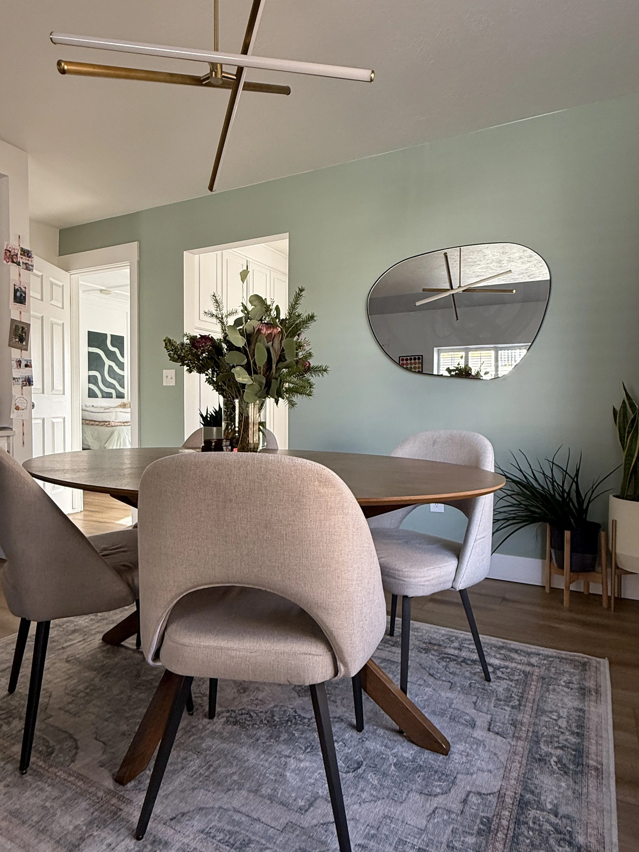 A round wooden dining table with four beige upholstered chairs in a room with light green walls, a modern abstract mirror, and a vase of flowers on the table. There is a patterned rug under the table, a potted plant to the right, and open doorways sh