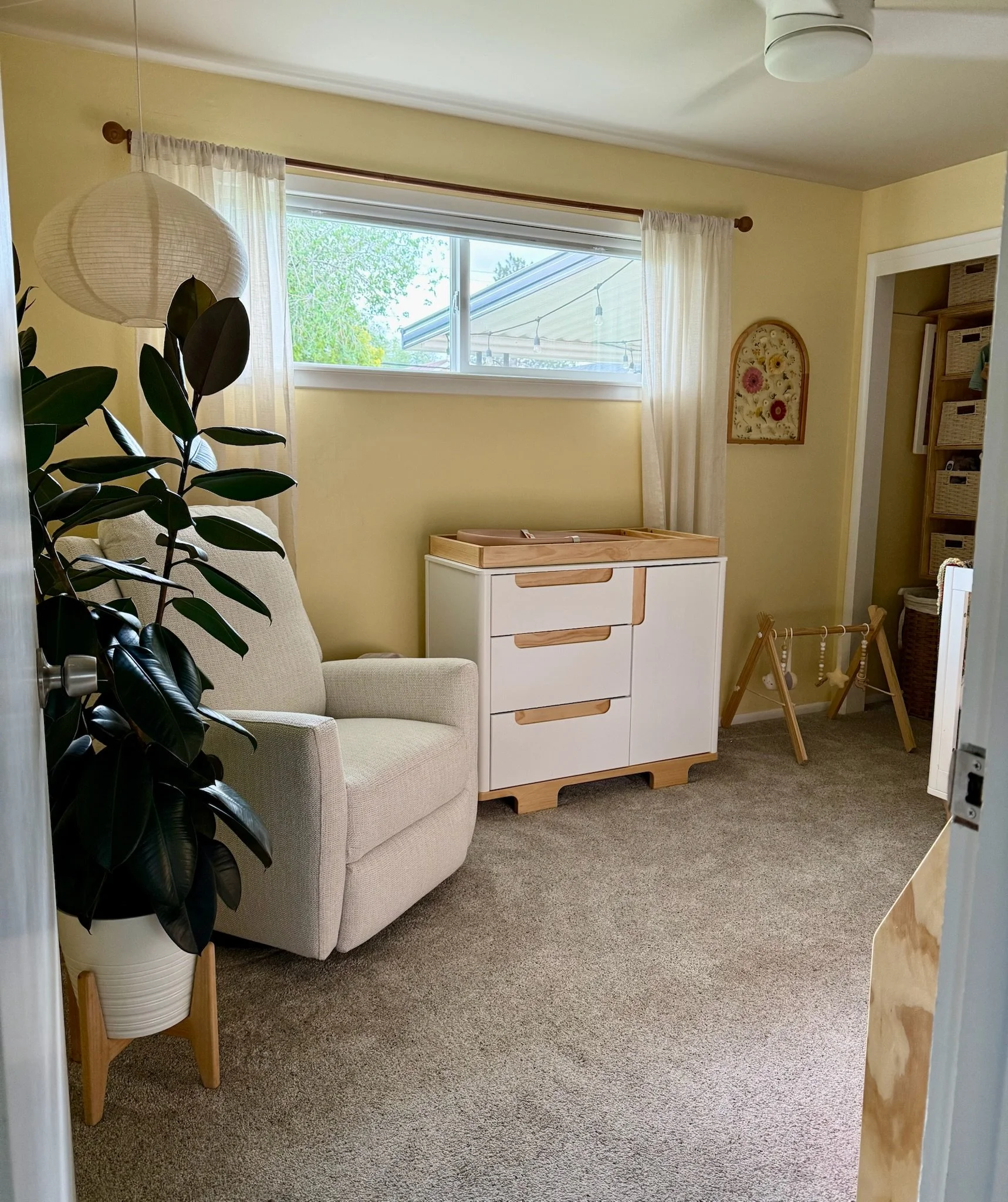 A cozy nursery corner with a beige armchair, a large potted rubber plant, a white storage cabinet with wooden accents, a window with white curtains, a small decorative wall hanging, and nearby shelves with storage baskets.
