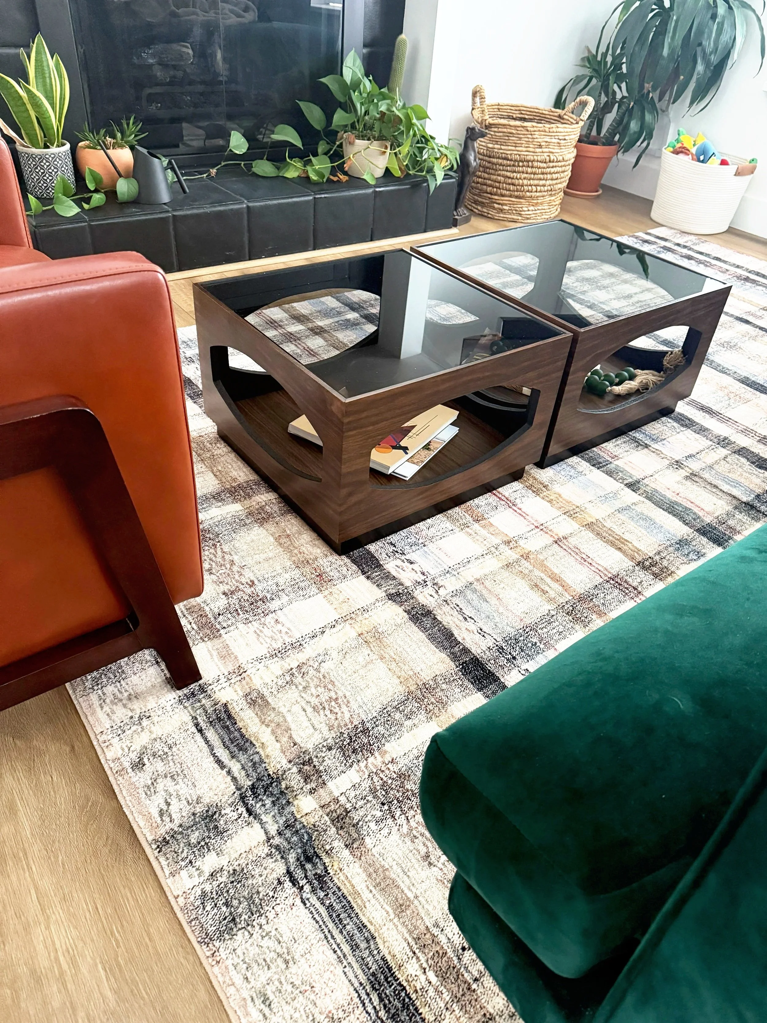 Living room with a glass-top coffee table, green velvet armchair, black fireplace mantel with potted plants and decorative items, and a colorful patterned area rug.