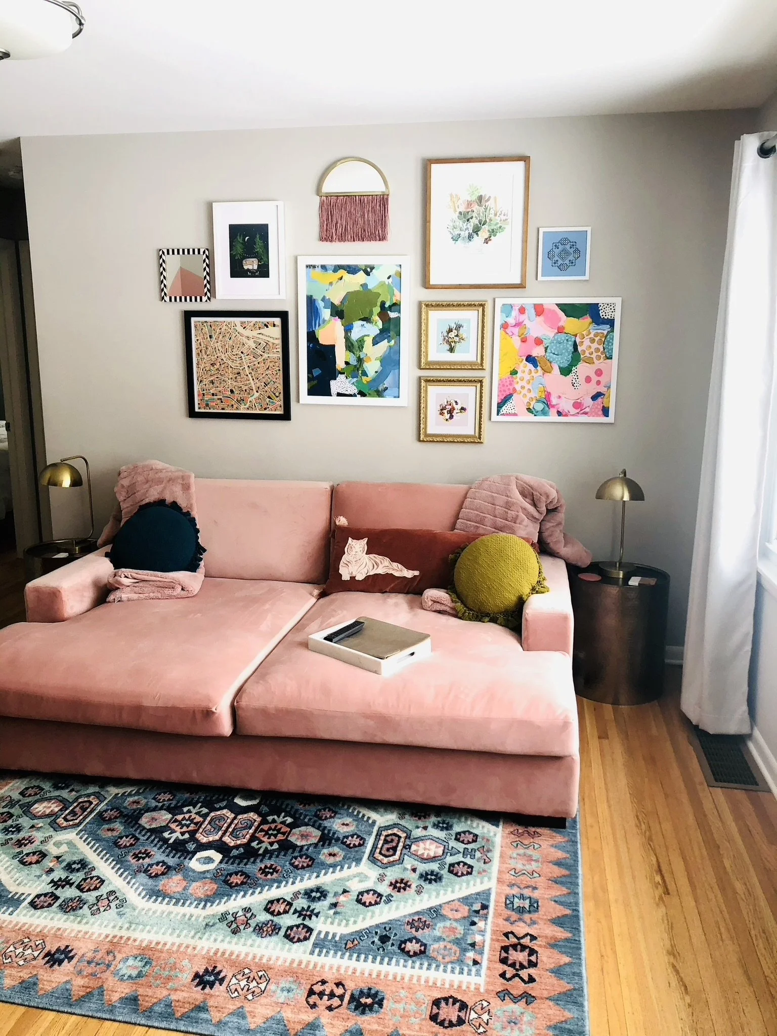 A cozy living room with a pink sectional sofa decorated with colorful pillows, including a dark blue one, a brown one with a cat illustration, and a round green textured pillow. A patterned area rug covers part of the hardwood floor. Several framed a
