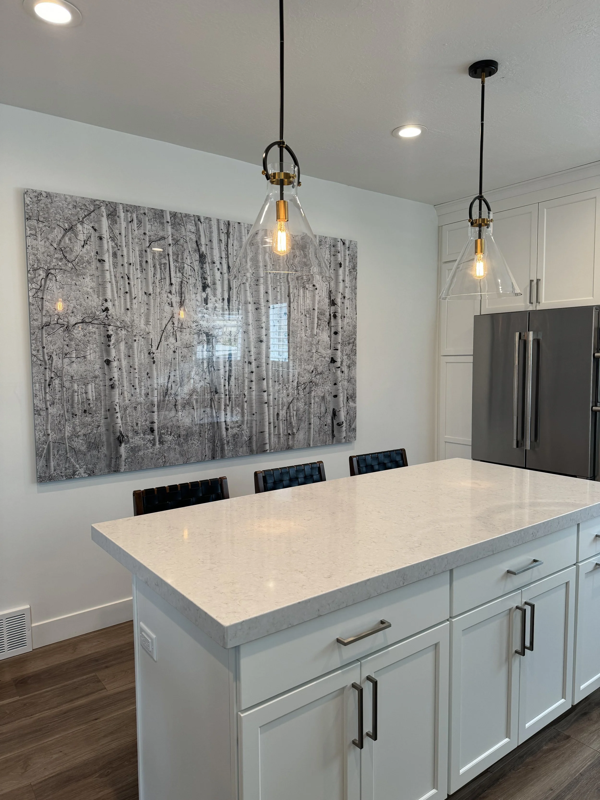 A kitchen with a white island counter, black cabinet handles, and a built-in oven. Two pendant lights hang from the ceiling above the counter. In the background, there's a large black and white artwork of a forest of birch trees.