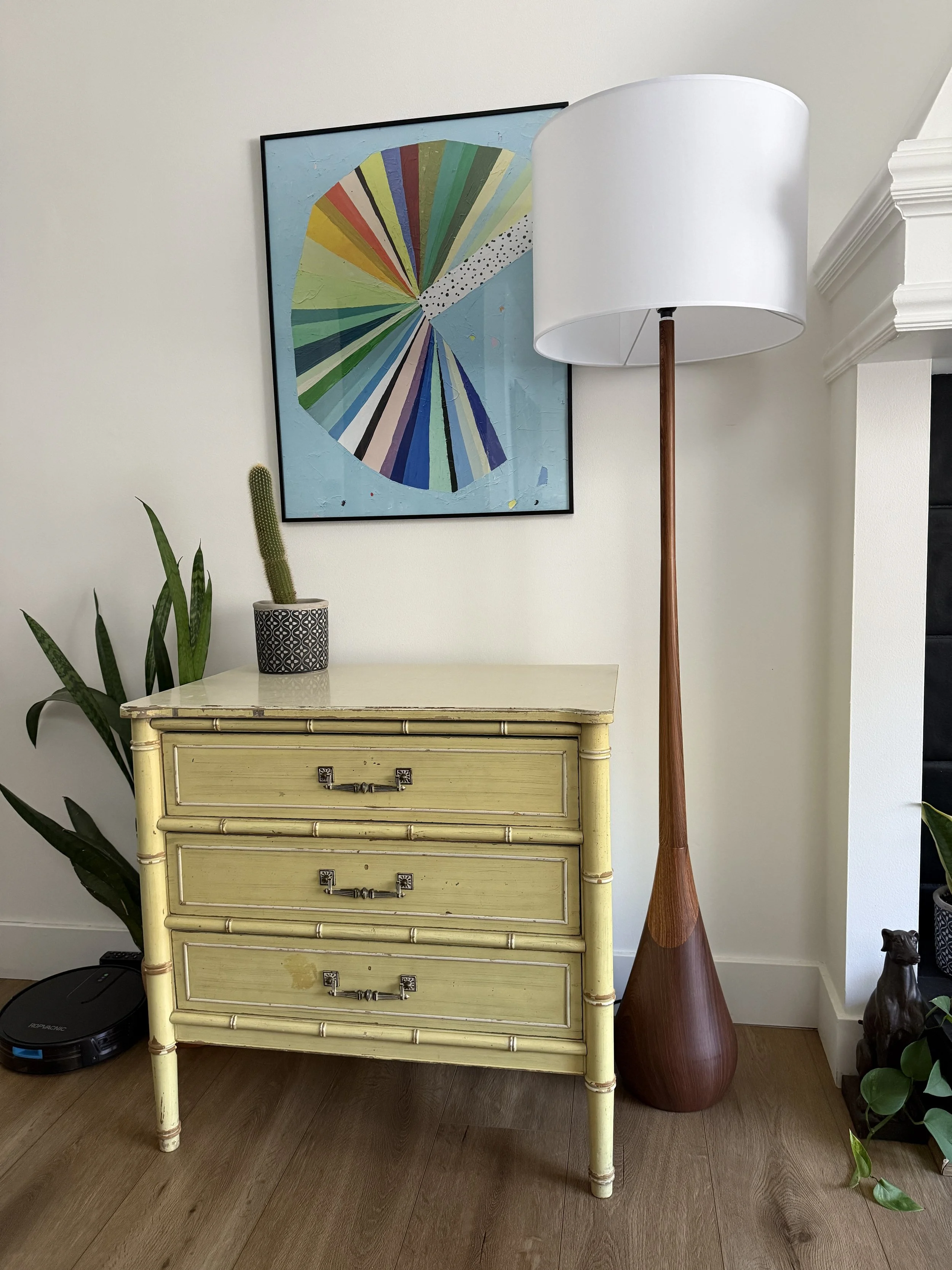 A yellow vintage dresser with four drawers and metallic handles, decorated with a potted cactus and greenery, positioned against a white wall with artwork, a tall wooden floor lamp, and a dark dog figurine nearby.
