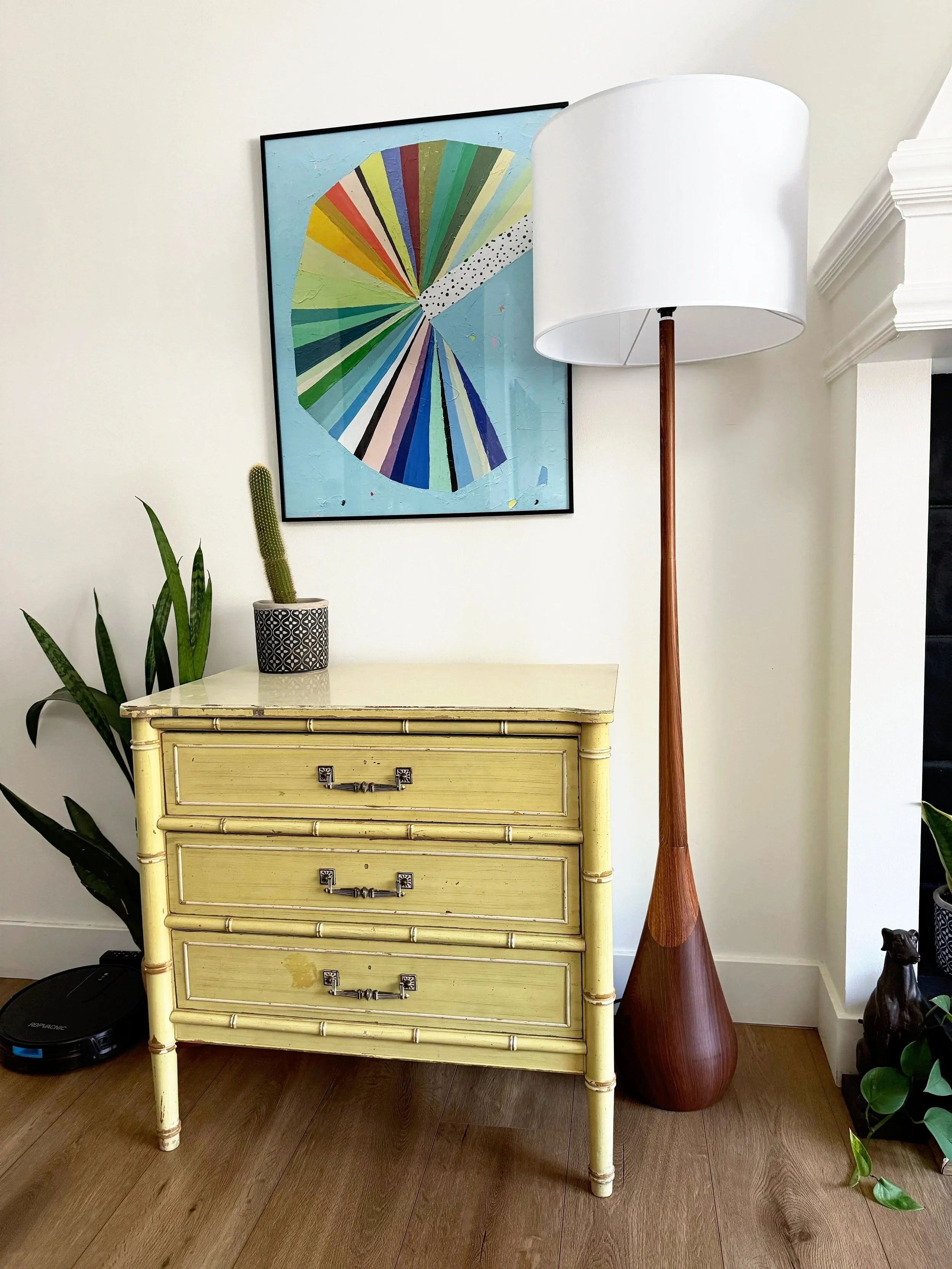 A yellow vintage dresser with three drawers and metal handles, topped with a potted cactus plant, is next to a tall wooden floor lamp with a white lampshade. There is a framed colorful geometric artwork on the wall behind the dresser, and some green plants on the right side of the image.