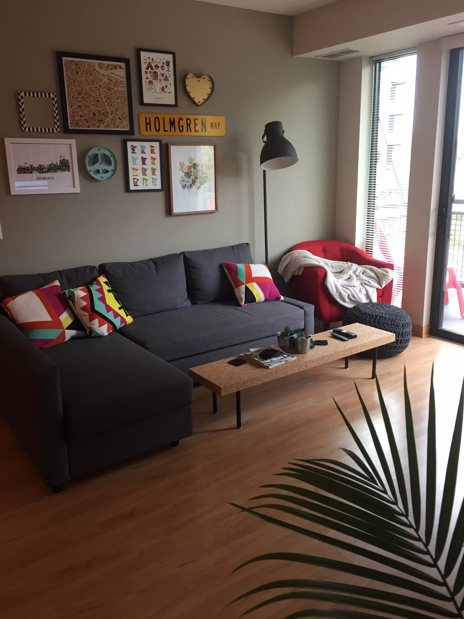 Living room with a gray sofa with colorful pillows, a red armchair with a blanket, a wooden coffee table with plants and remote controls, a large window with vertical blinds, a black floor lamp, and wall decorations including framed pictures, a map, 